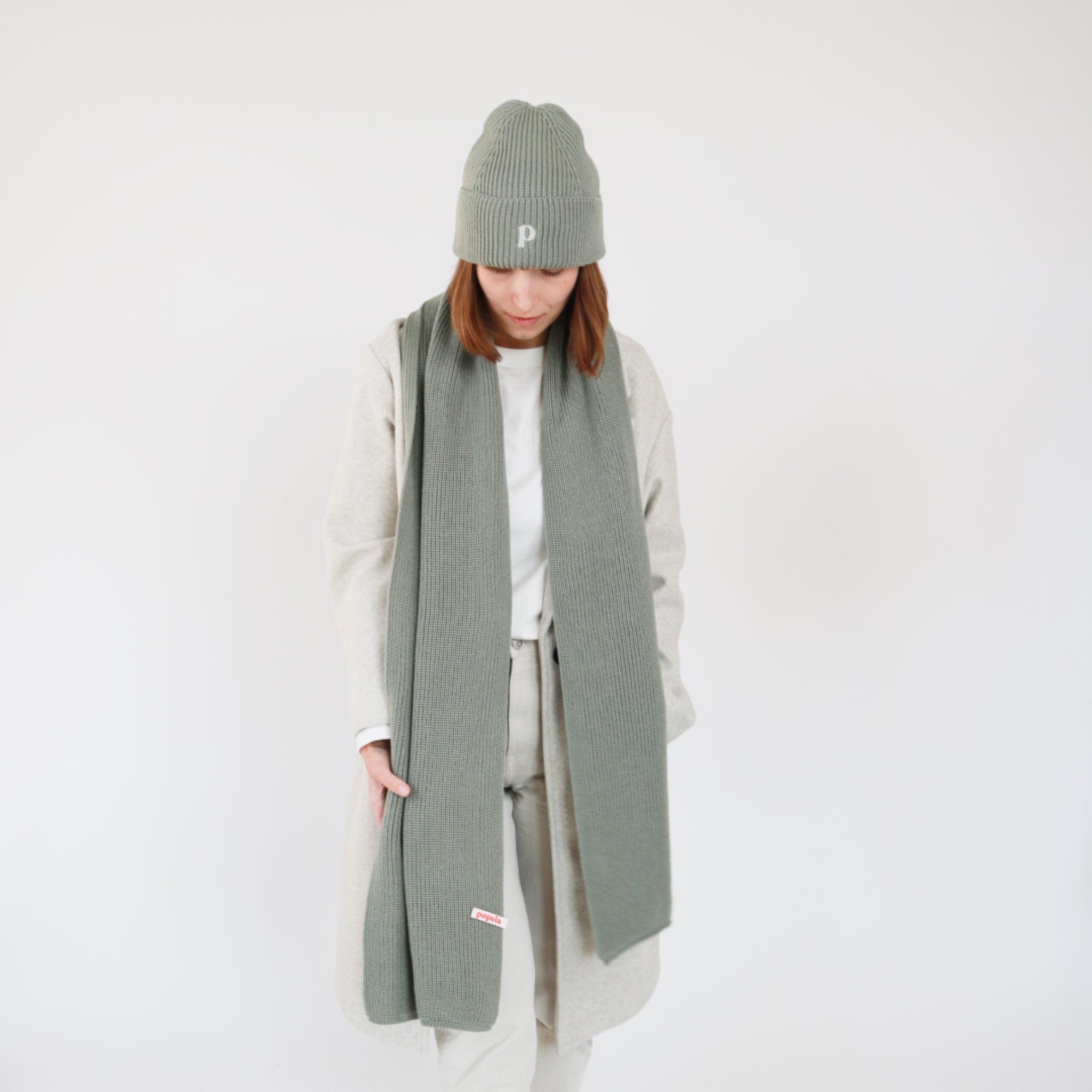 Eine Person trägt einen langen, grünen Schal und eine passende grüne Mütze über einem hellen Mantel.