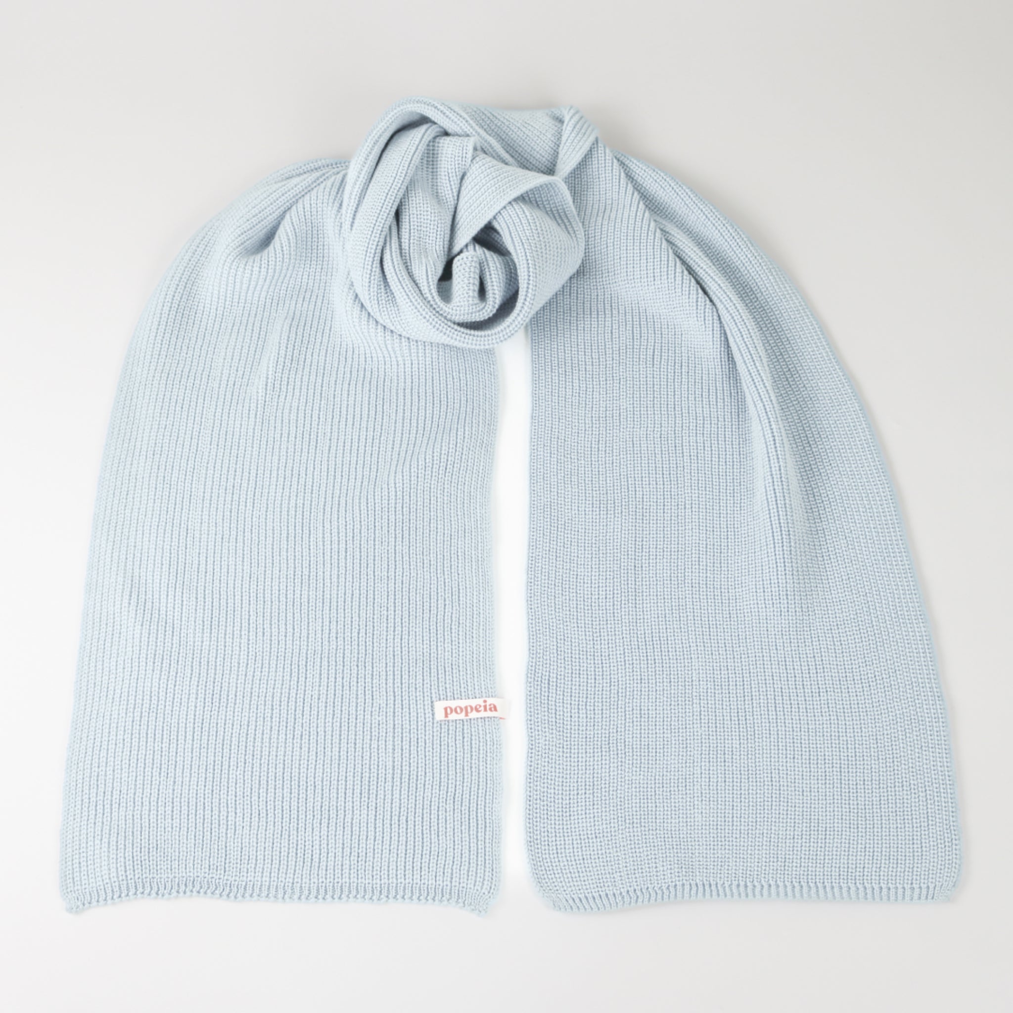 Hellblauer Strickschal mit Etikett „popeia“, sorgfältig zusammengelegt auf weißem Hintergrund.