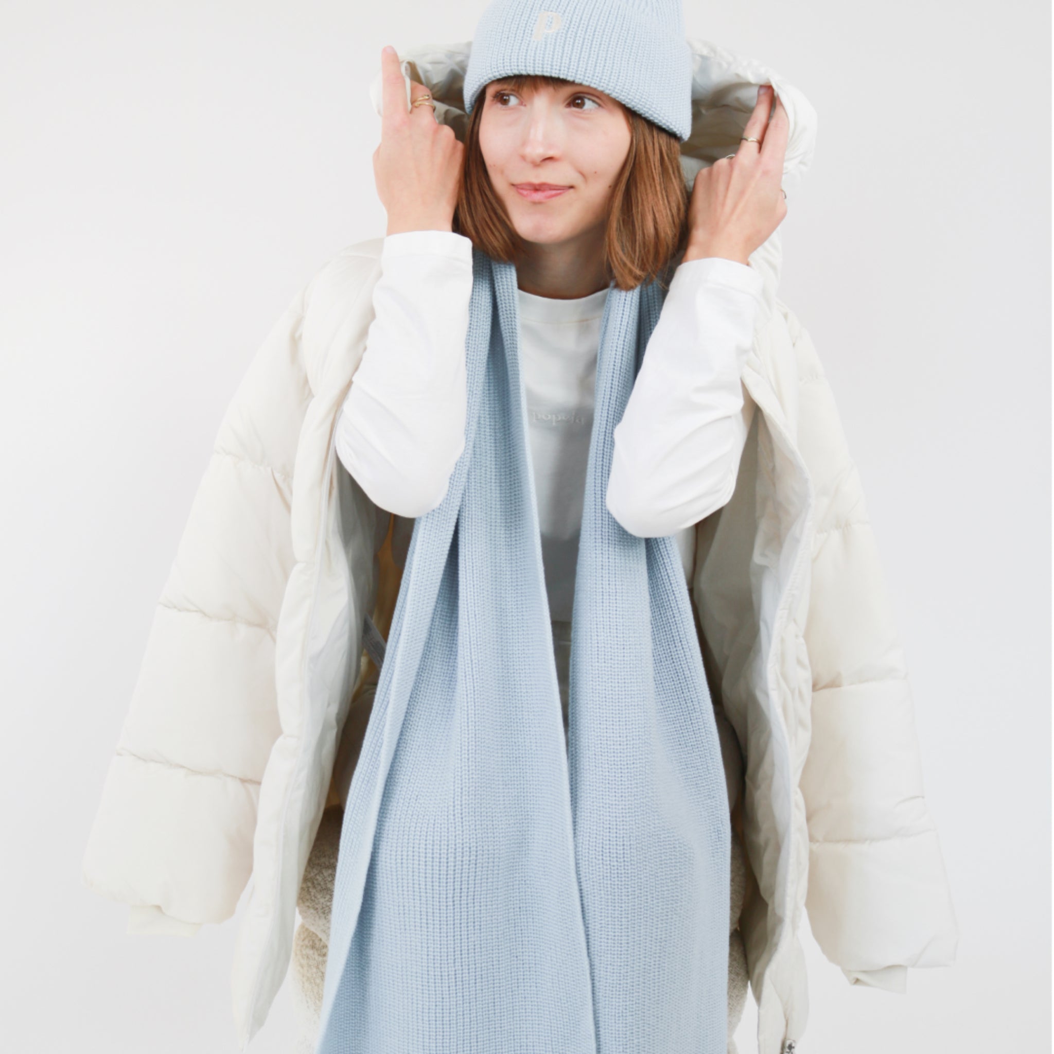 Frau in blauer Strickmütze und Schal trägt weißen Wintermantel mit Kapuze, vor weißem Hintergrund.