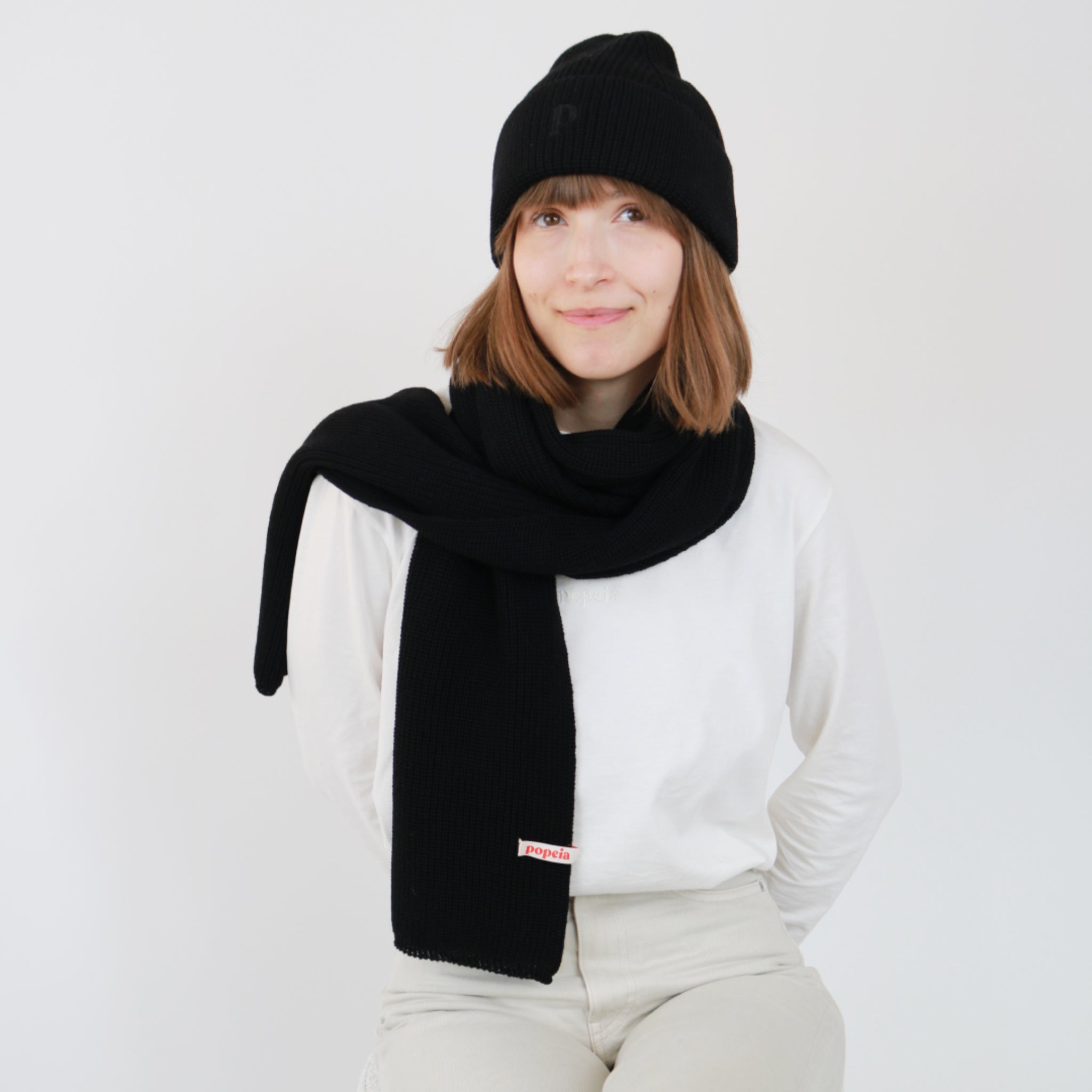 Eine Person trägt ein schwarzes Mützchen und einen passenden Schal über einem weißen Pullover vor einem neutralen Hintergrund.