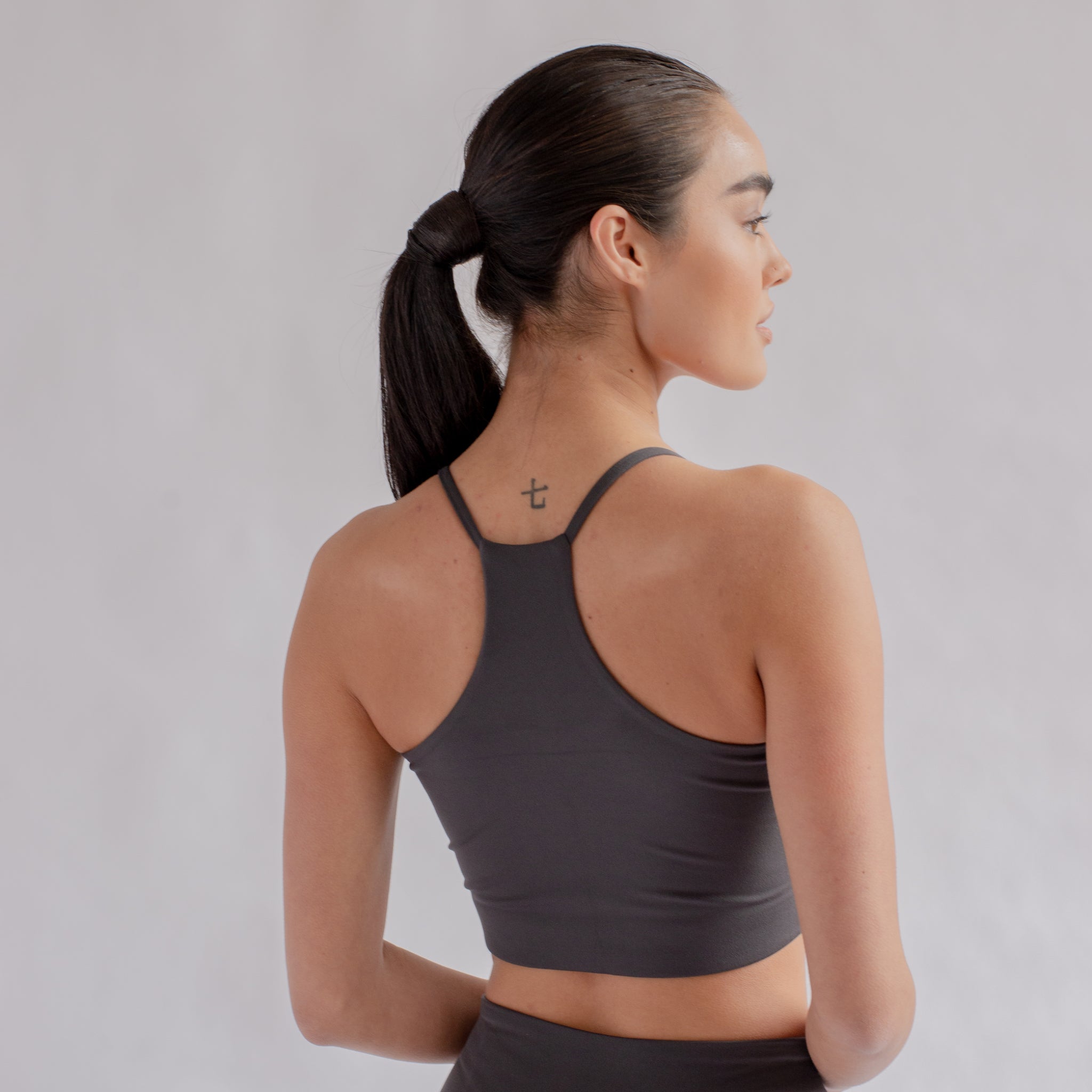 Frau in dunkelgrauem Sporttop mit Pferdeschwanz steht seitlich, zeigt ein Tattoo auf dem Nacken, vor hellem Hintergrund.