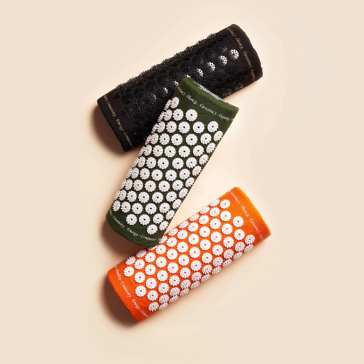 Drei Akupressur-Matten in Schwarz, Grün und Orange mit weißen Stachelmustern liegen auf einem beigen Hintergrund.