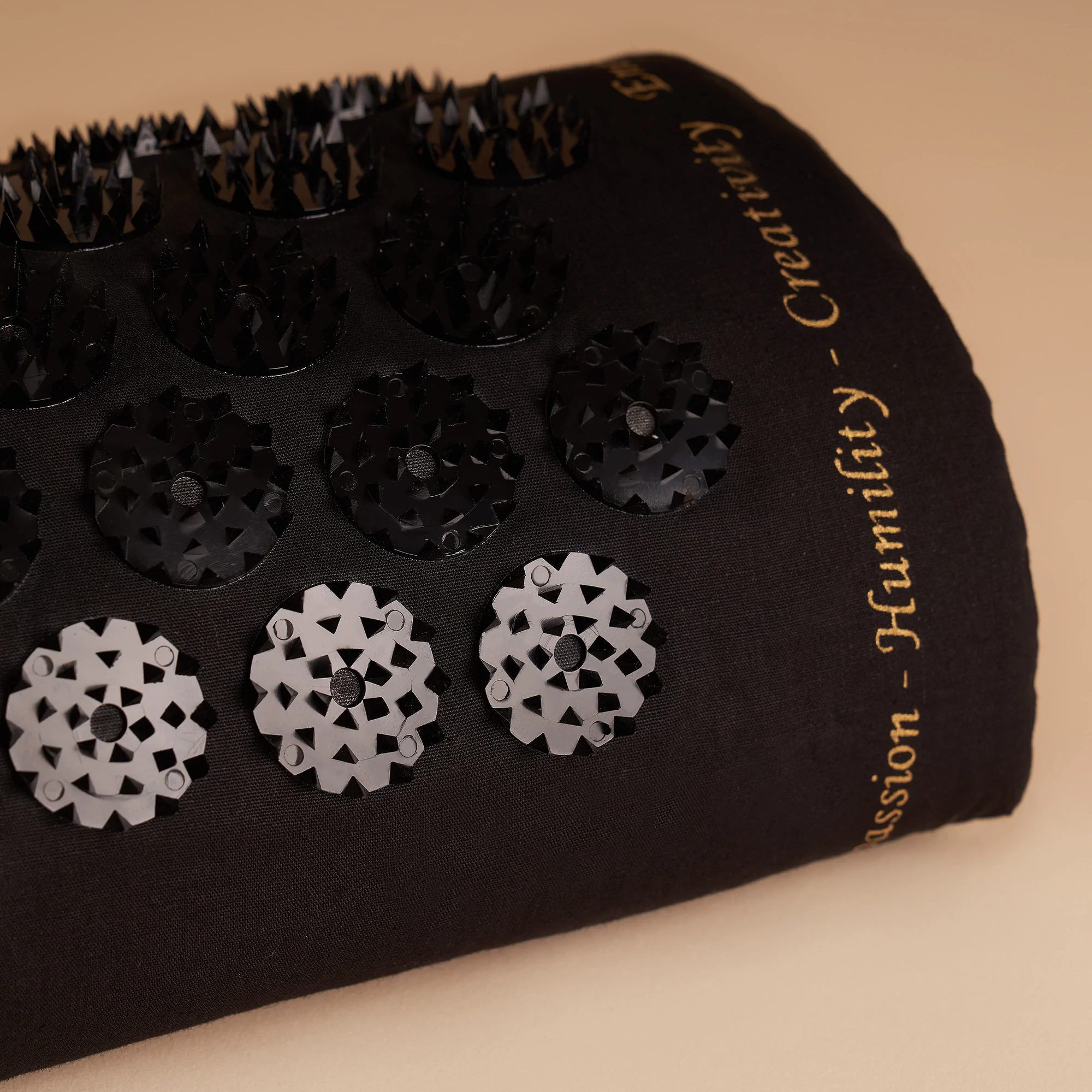 Schwarzes Nackenkissen mit kleinen runden Akupressur-Spitzen und goldener Aufschrift auf beigem Hintergrund.