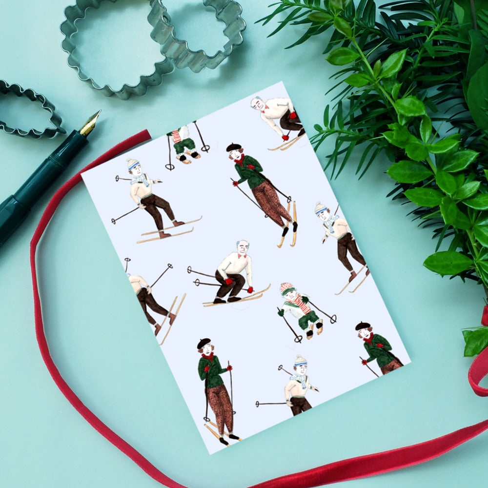 Karte mit Skifahrern vor hellem Hintergrund, umgeben von Pflanzen, Ausstechformen, Stift und rotem Band.