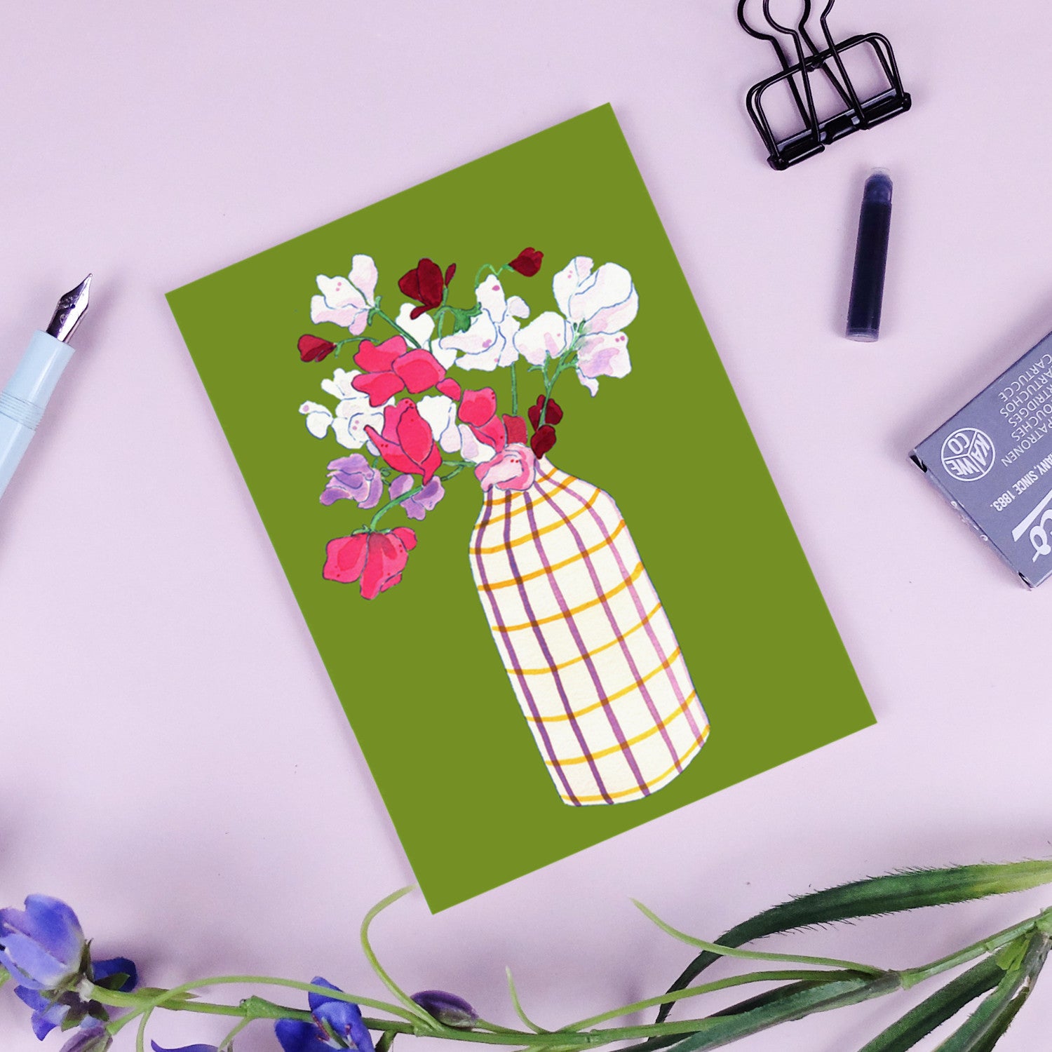 Karte mit Blumen in kariertem Vasenmotiv auf grünem Hintergrund, umgeben von Schreibutensilien auf rosa Tisch.