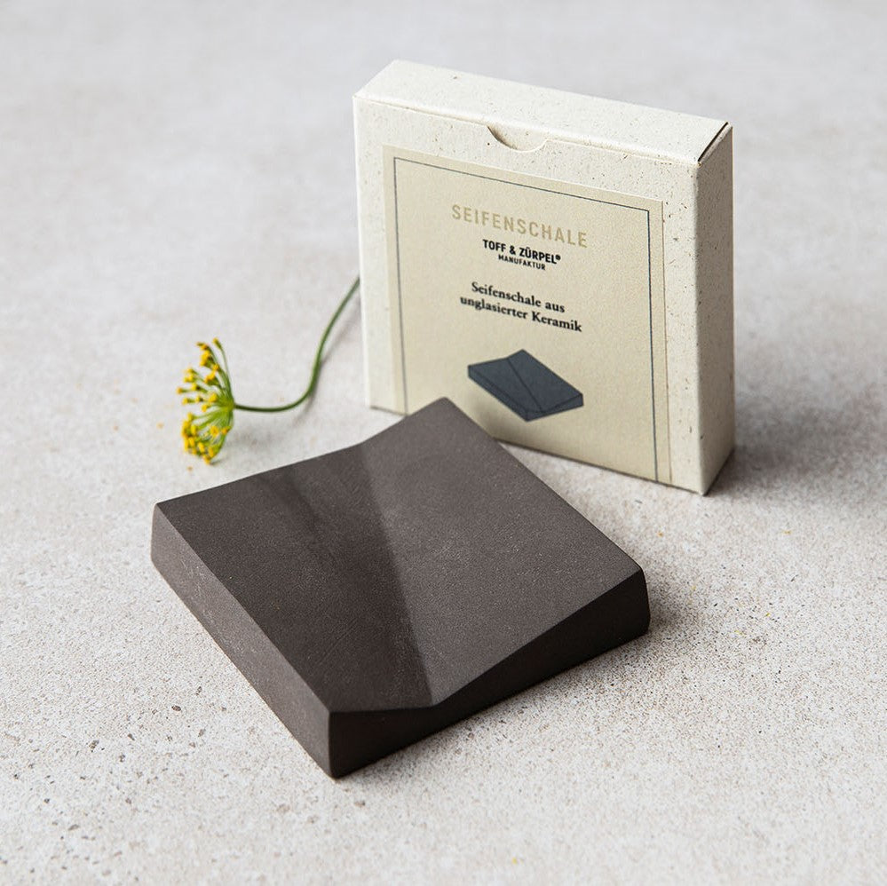 Dunkle, unglasierte Keramikseifenschale vor ihrer Verpackung, neben einer gelben Blume auf grauem Hintergrund.
