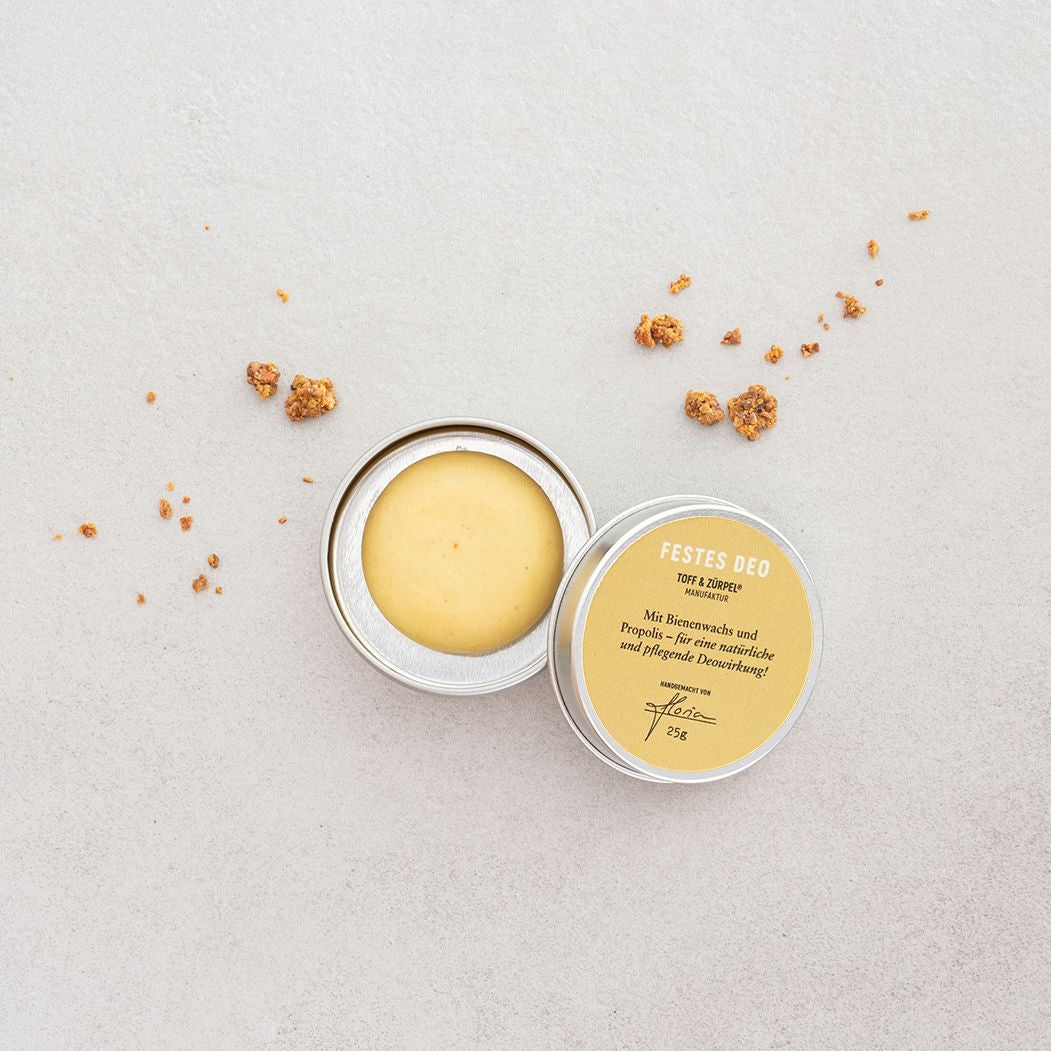 Runde Dose mit festem Deodorant auf hellem Hintergrund, daneben liegen kleine braune Krümel verstreut.