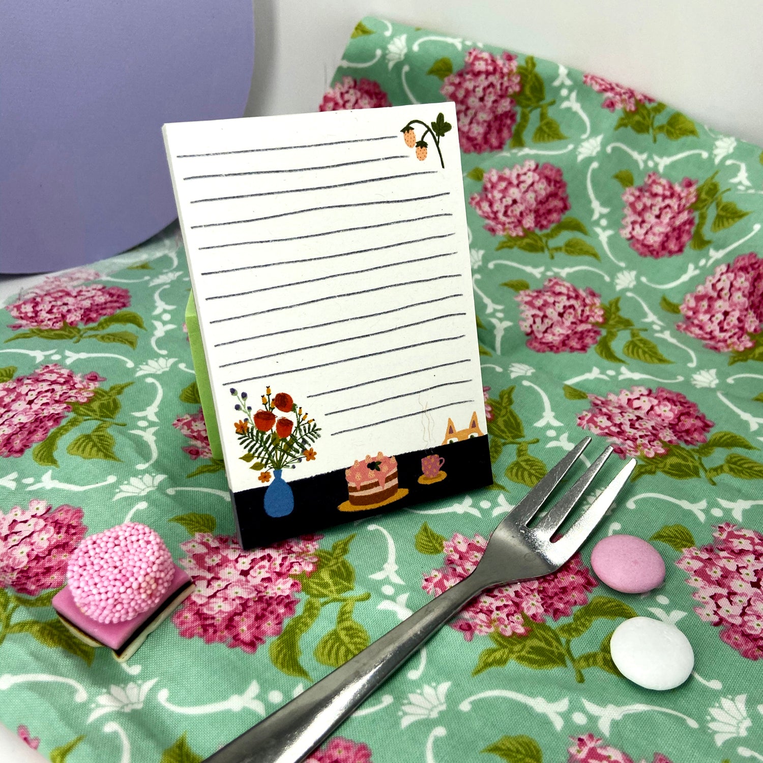 Notizblock mit Zeichnungen von Blumen und Kuchen auf gemustertem Stoff, daneben eine Gabel und bunte Süßigkeiten.