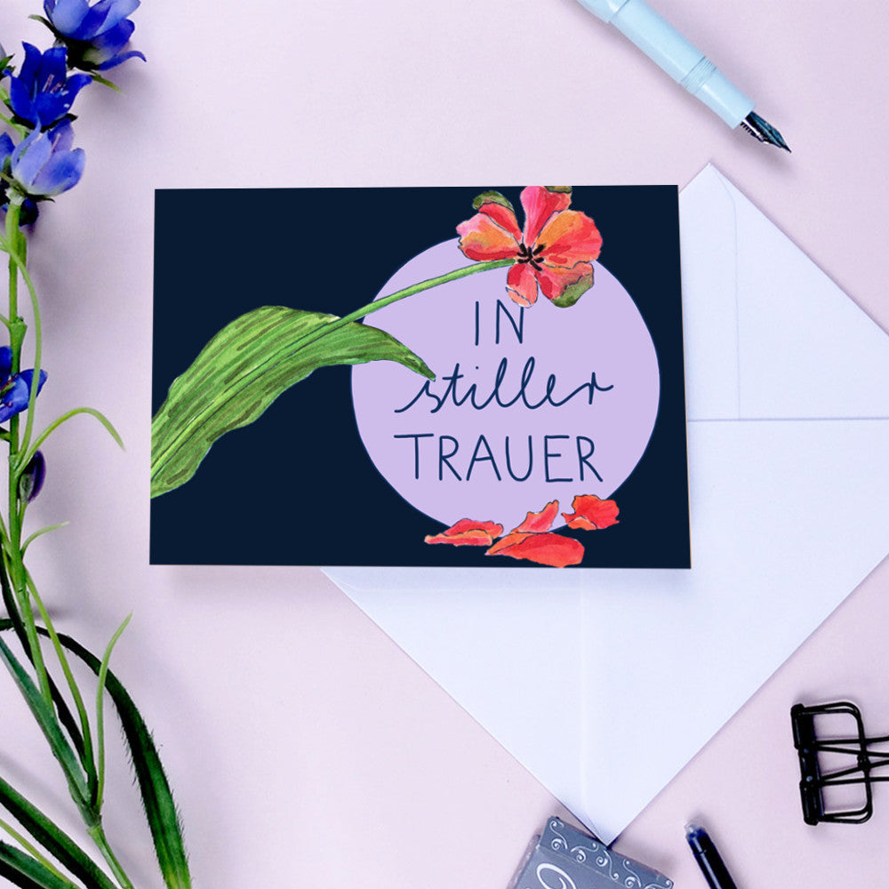 Trauerkarte mit Blumenmotiv und Text "In stiller Trauer" auf einem lilafarbenen Kreis, umgeben von Schreibutensilien.