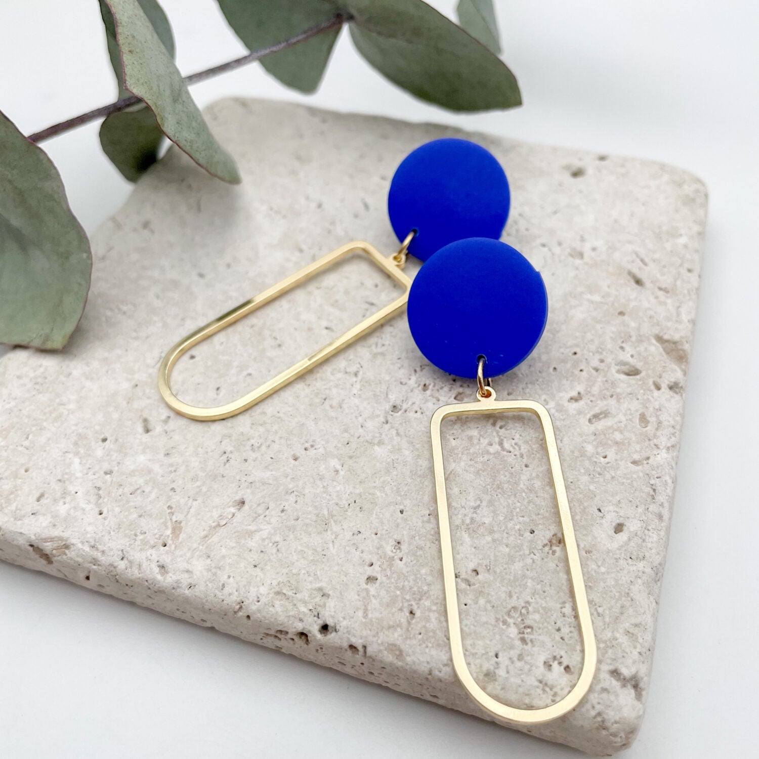 Goldene Ohrhänger mit blauen runden Oberteilen, auf Steinplatte mit grünen Blättern dekoriert.