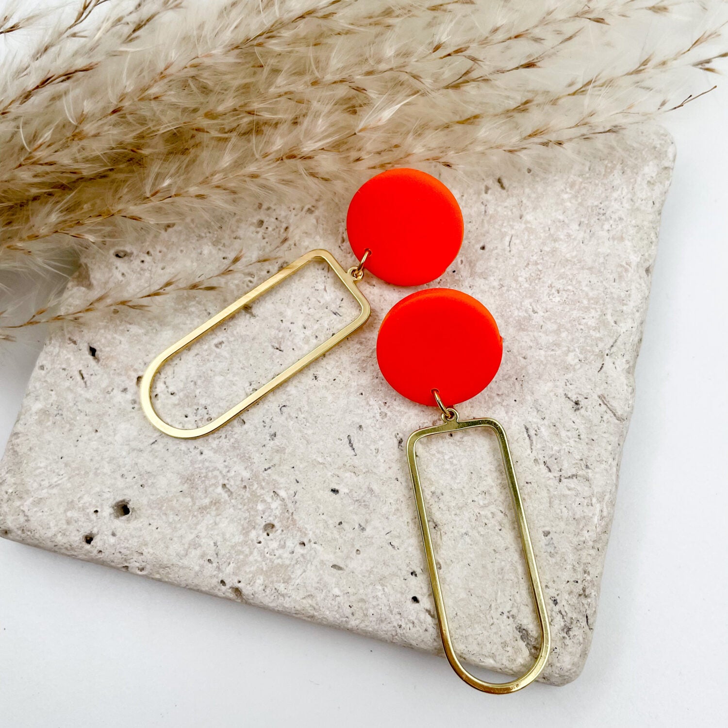 Goldene Ohrringe mit roten Kreisanhängern auf betonähnlicher Oberfläche, dekoriert mit getrockneten Gräsern.