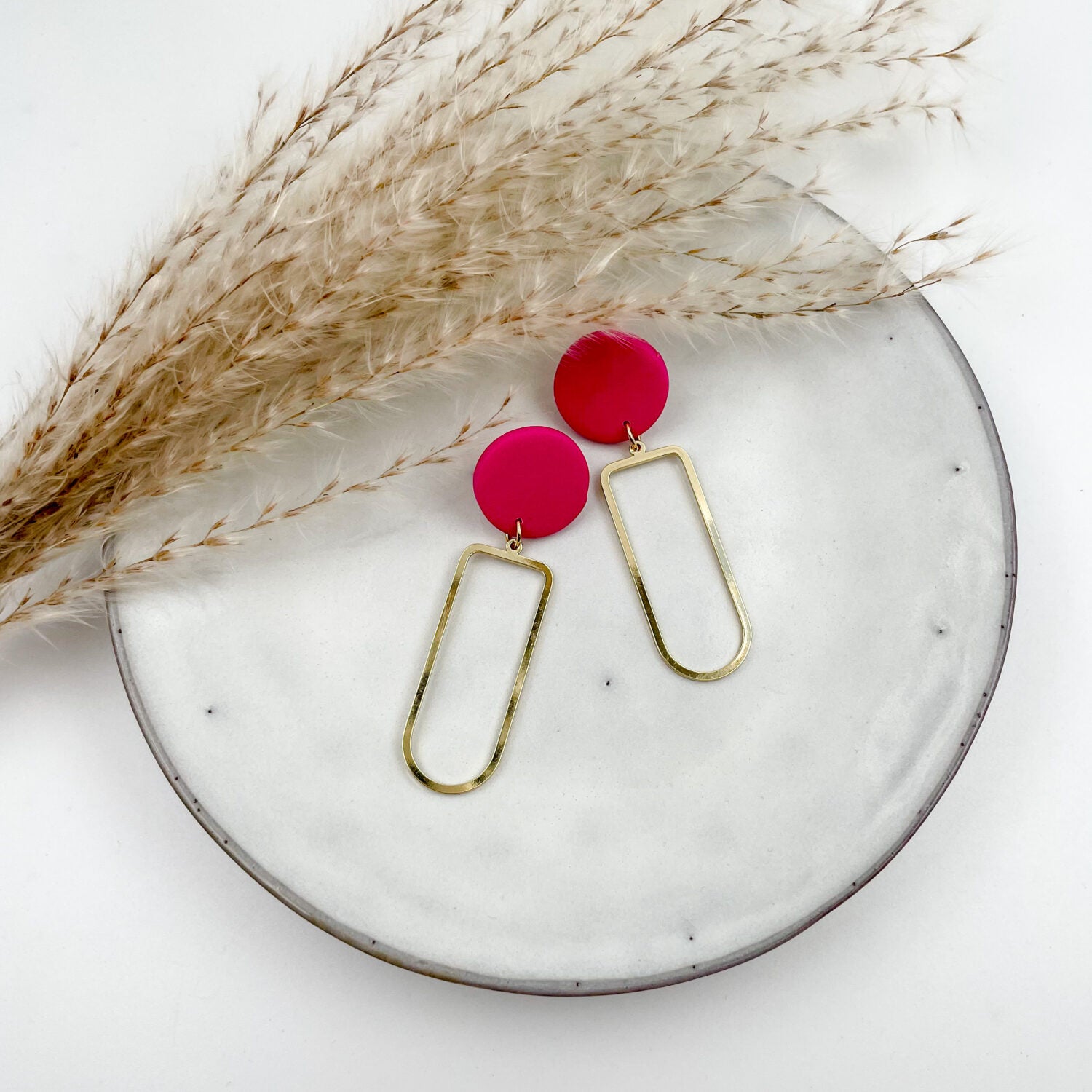 Goldene Ohrringe mit pinken Kreisen auf weißem Teller, neben getrocknetem Pampasgras arrangiert.