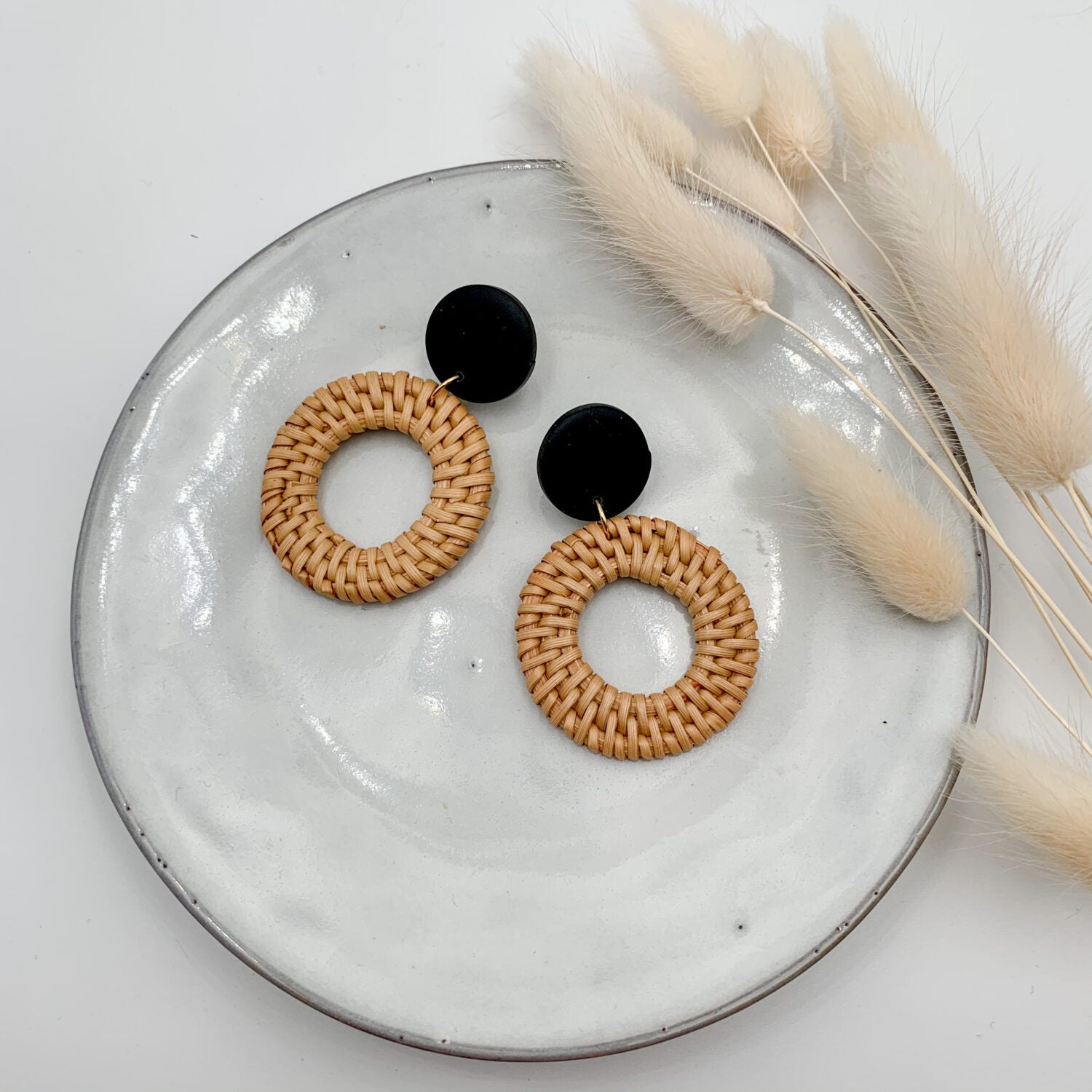 Zwei Ohrringe aus Rattan und schwarzem Material auf einem weißen Teller mit getrockneten Gräsern.