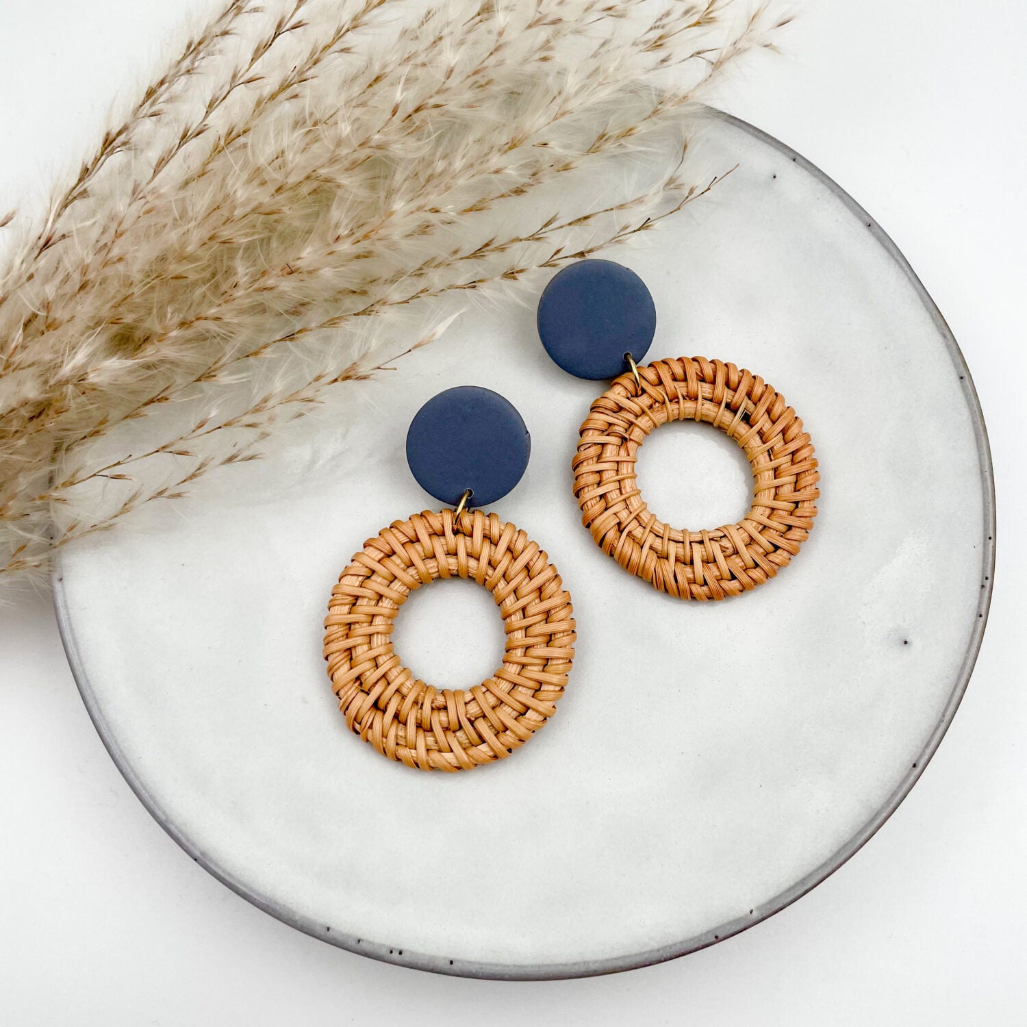 Runde handgeflochtene Ohrringe mit blauen Akzenten liegen auf einem Teller neben getrocknetem Pampasgras.