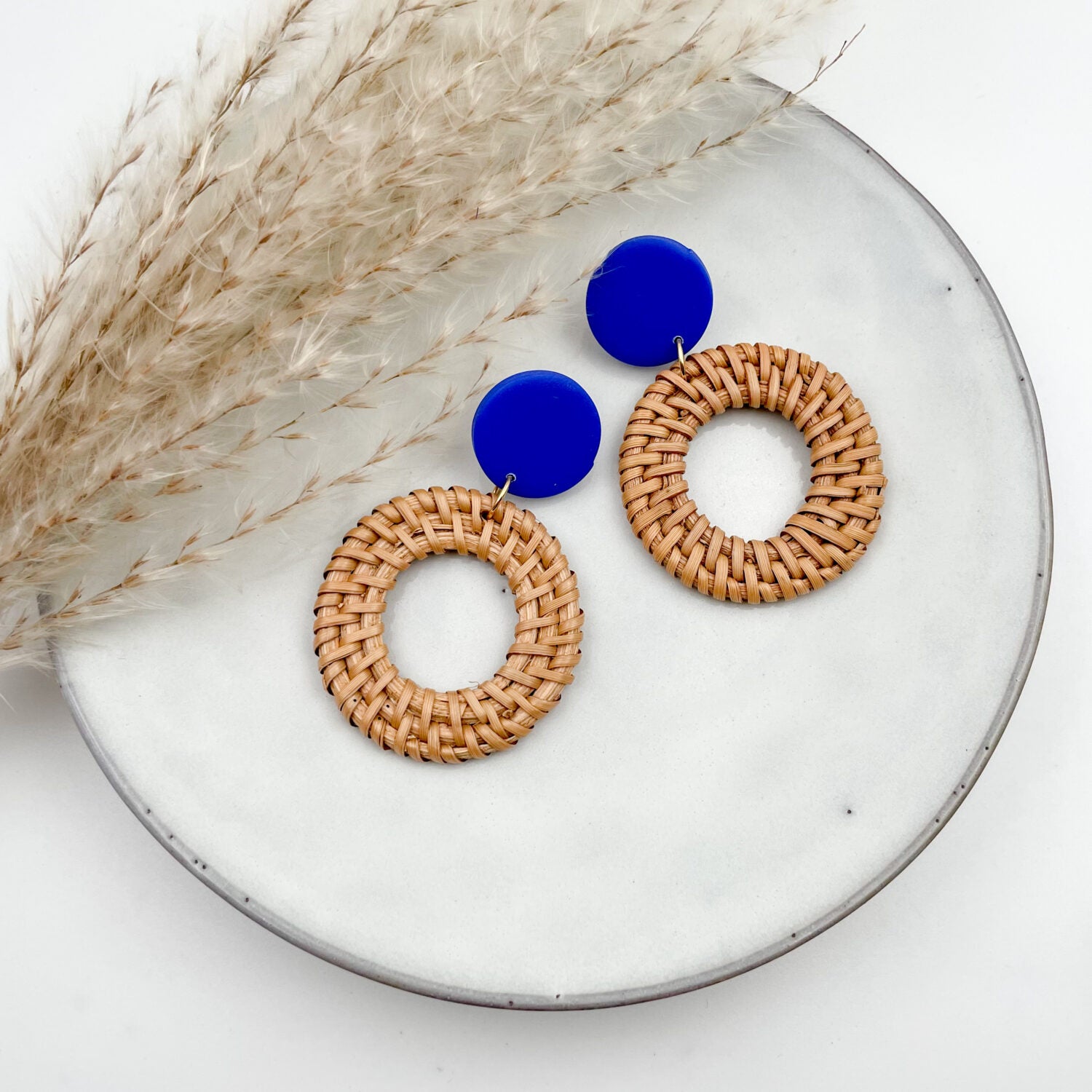 Runde Ohrringe aus Rattan mit blauen Anhängern auf einem weißen Teller neben getrockneten Gräsern.