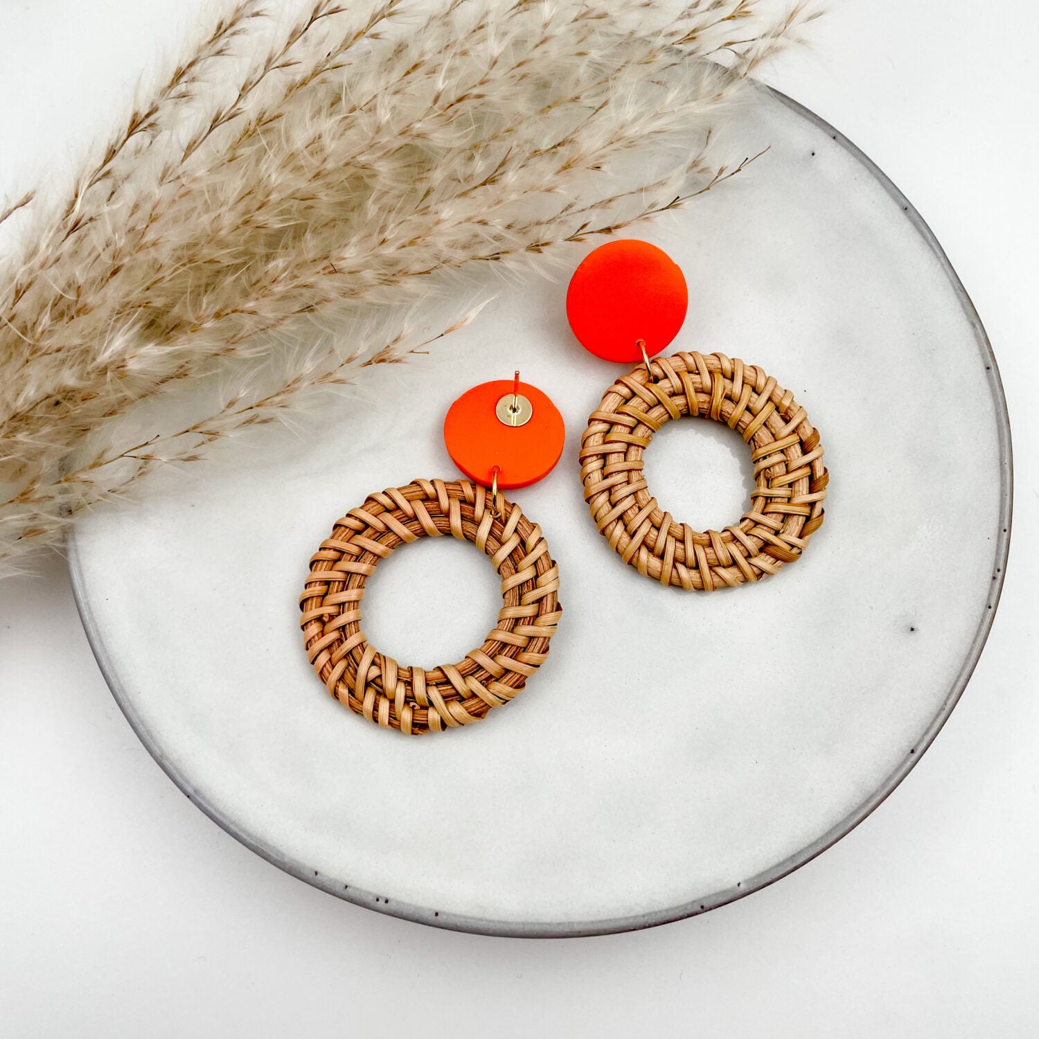 Zwei geflochtene Ohrringe mit orangefarbenem Akzent auf einem weißen Teller neben getrockneten Halmen.