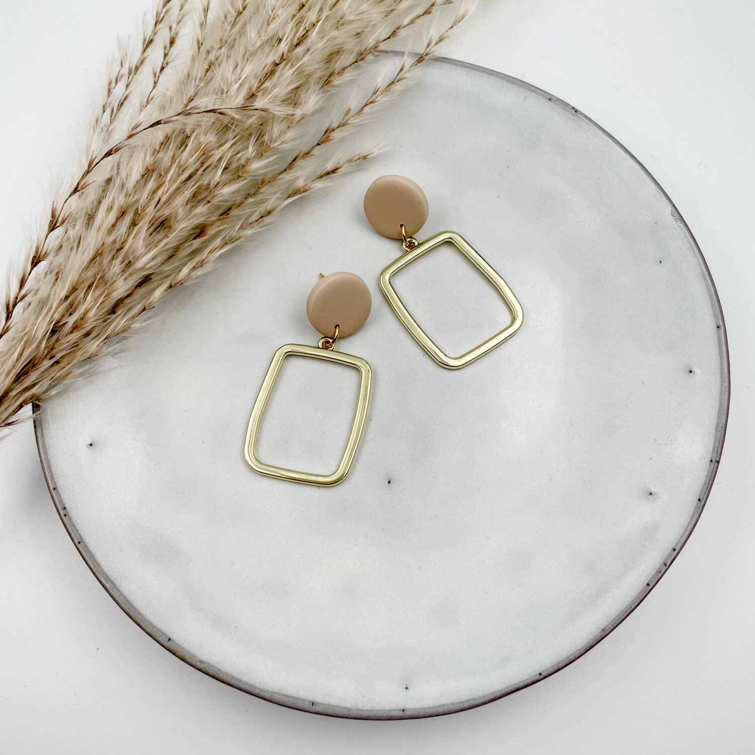 Goldene Ohrringe mit beige Kreolen liegen auf einem weißen Teller neben getrocknetem Pampasgras.