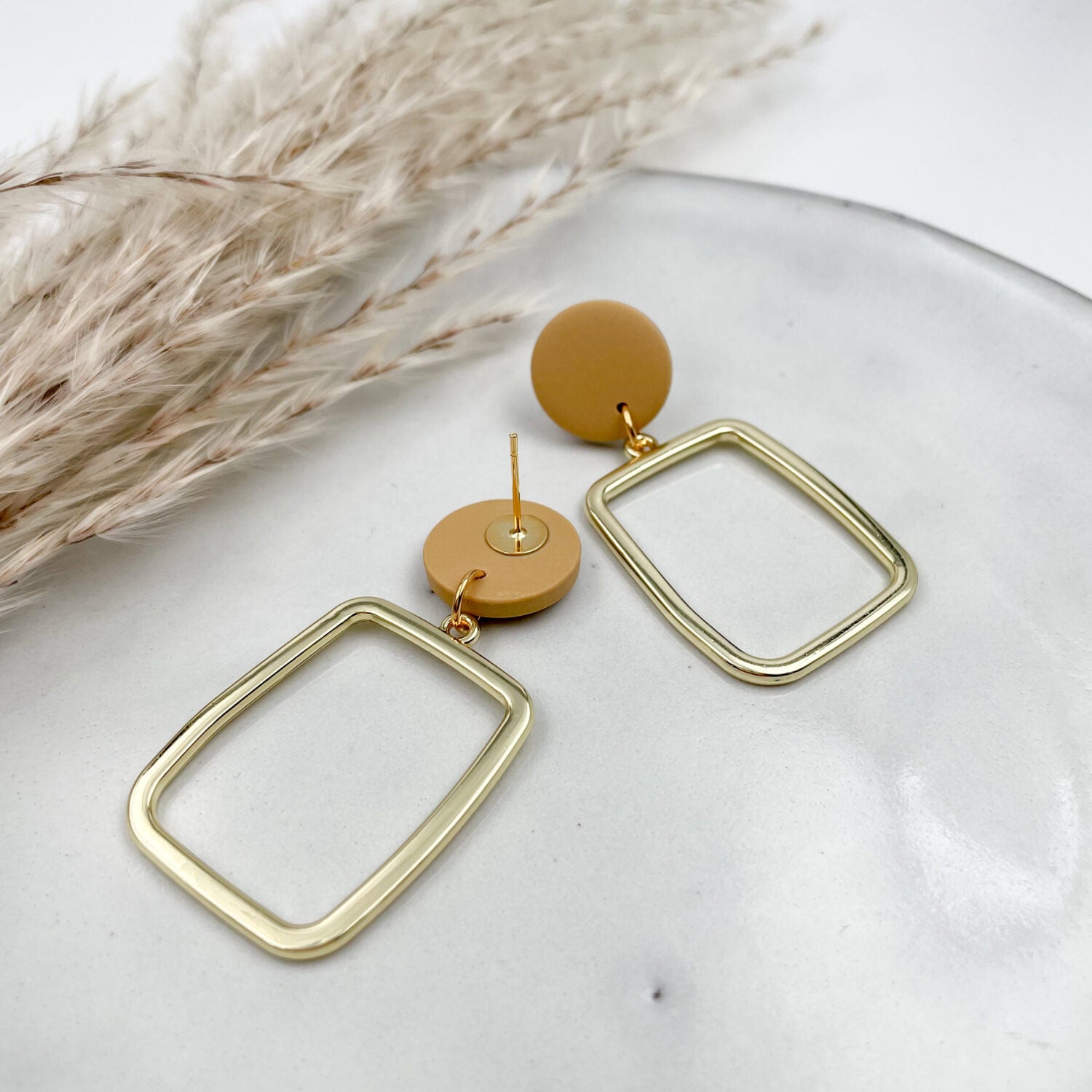 Goldfarbene Ohrringe mit rechteckigem Anhänger liegen auf einem weißen Teller neben getrocknetem Pampasgras.