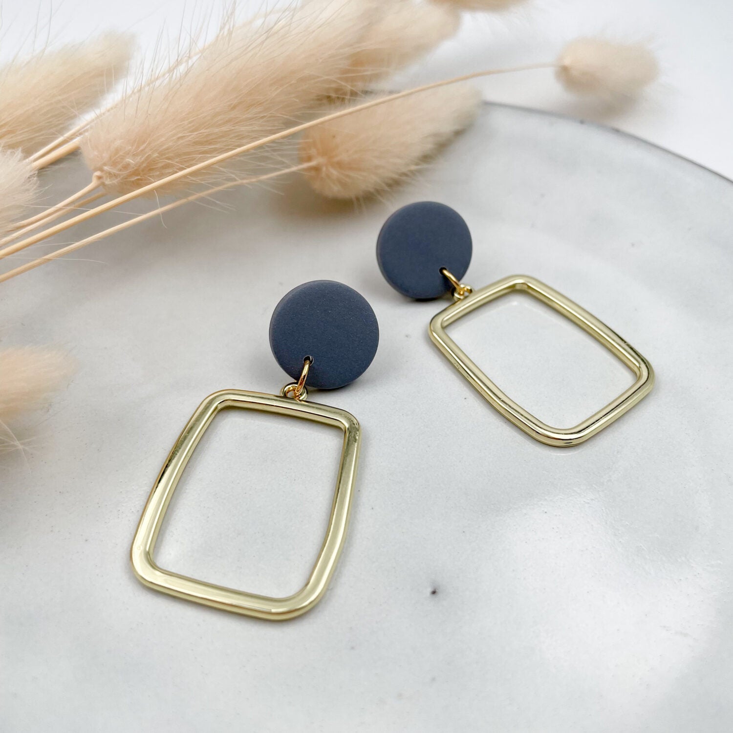 Goldene Ohrringe mit blauem Kreis und rechteckigem Anhänger auf grauem Teller, daneben beige Trockenblumen.