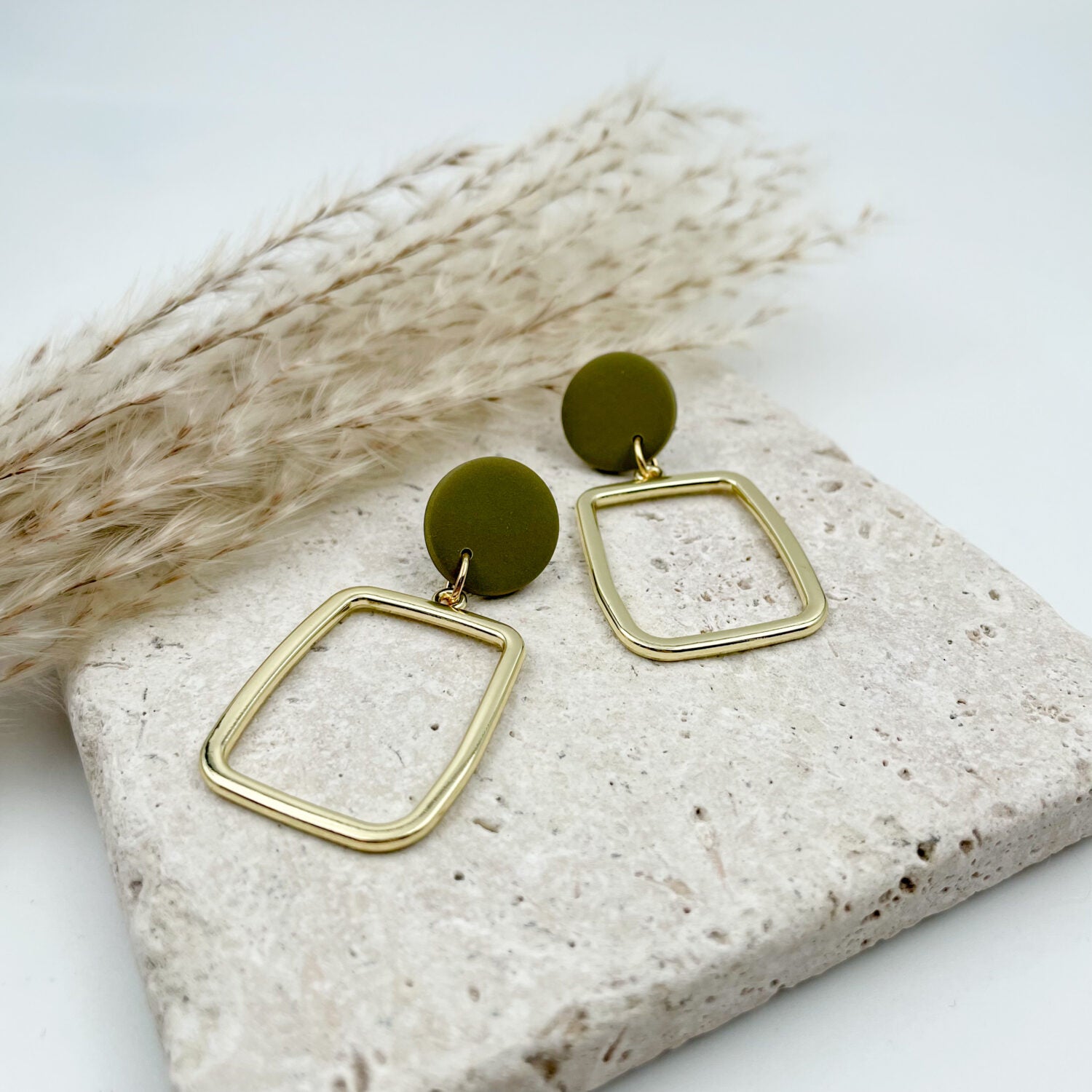 Goldene Ohrringe mit olivgrünen Kreisen auf einer Steinplatte, neben Getreideähren liegend.