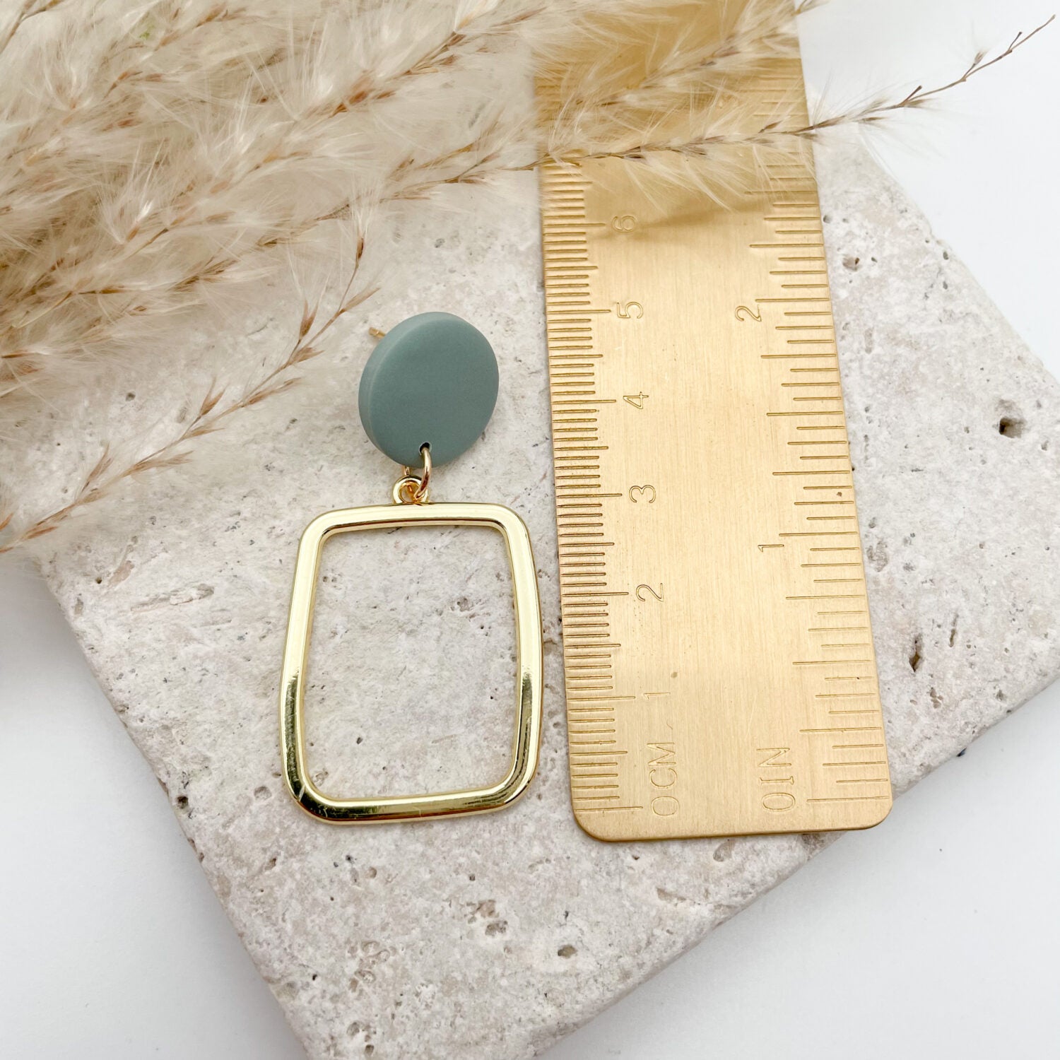 Goldener Ohrring mit grünem Kreis neben einem Lineal auf einer Steinoberfläche, umgeben von zarten Gräsern.