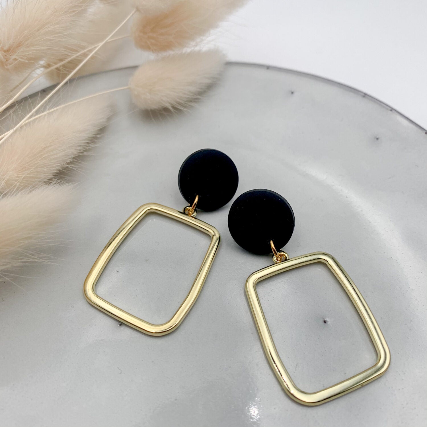 Goldene und schwarze Ohrringe auf einem weißen Teller, daneben liegen beige Trockenblumen.