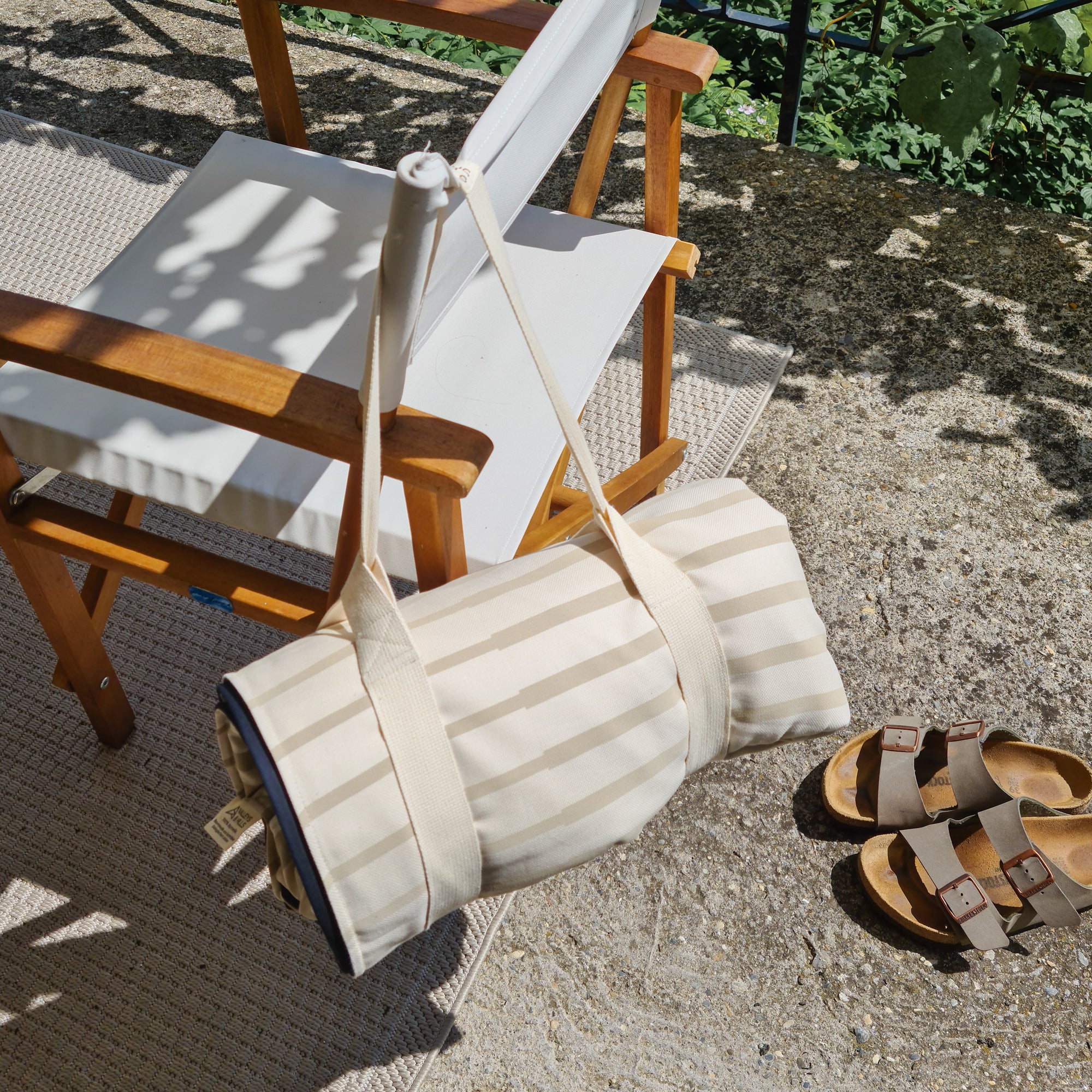 Beiger Klappstuhl und gerollte Decke auf einer Terrasse, daneben ein Paar Sandalen auf dem Boden.