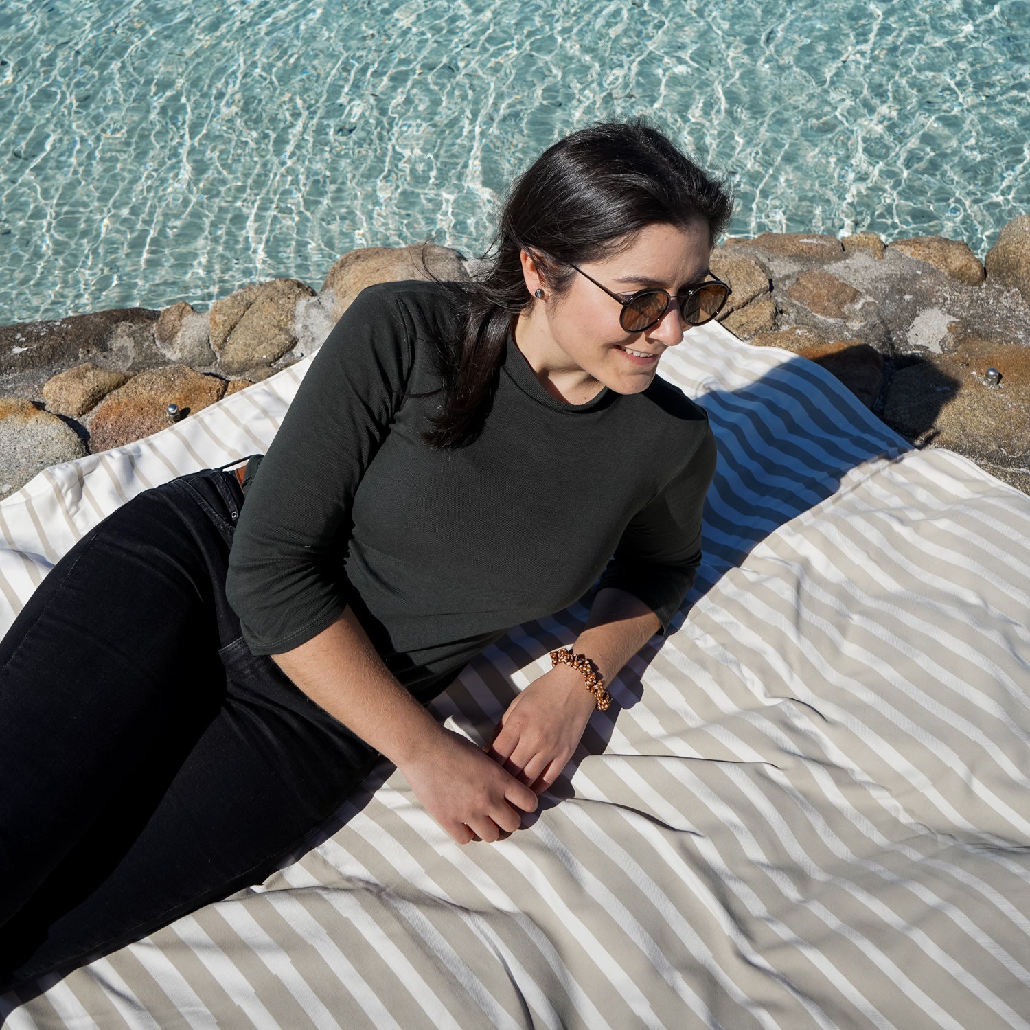 Frau in Sonnenbrille liegt auf gestreiftem Handtuch am Rand eines klaren Pools.
