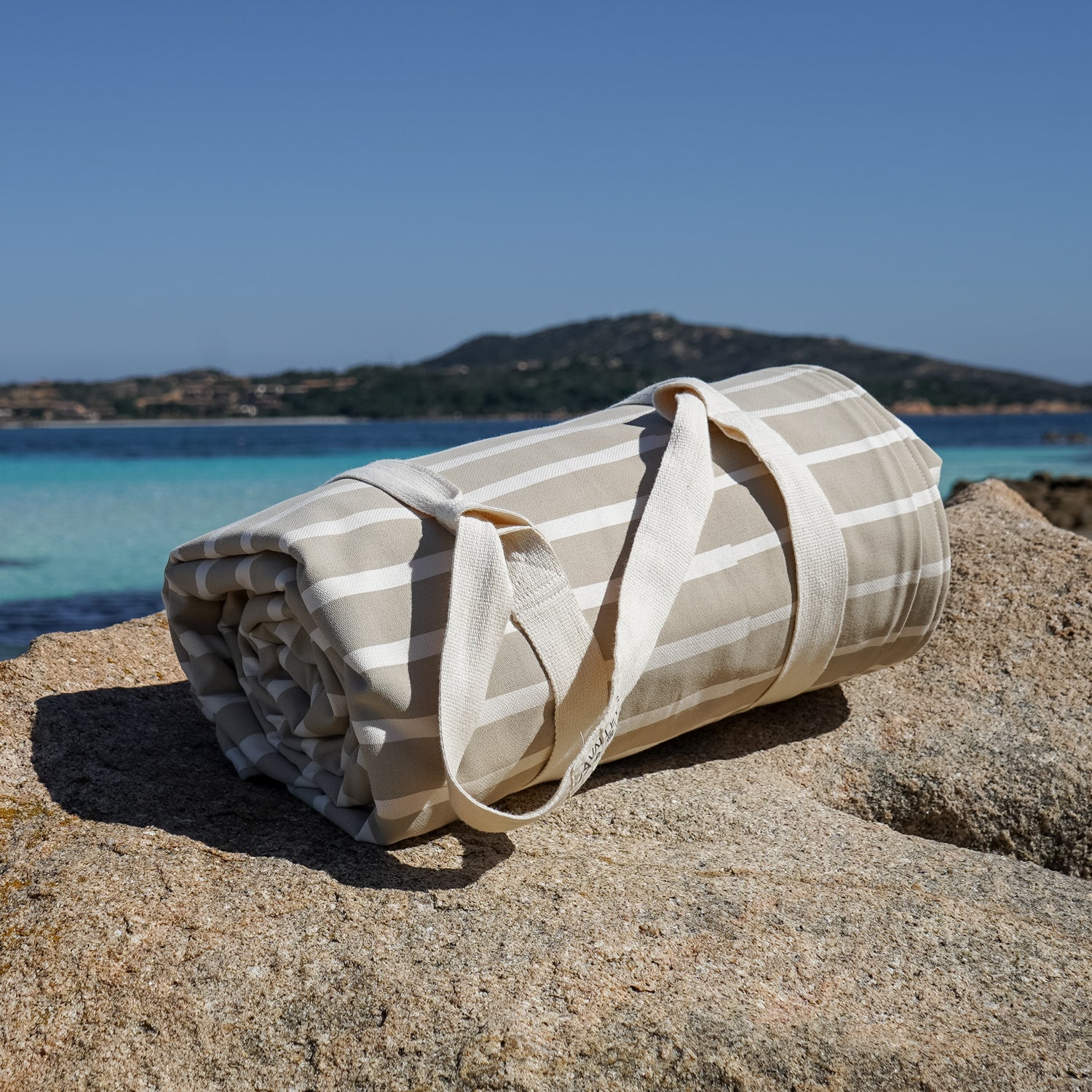 Gerollte gestreifte Strandmatte auf Felsen vor türkisblauem Meer und Hügel im Hintergrund.
