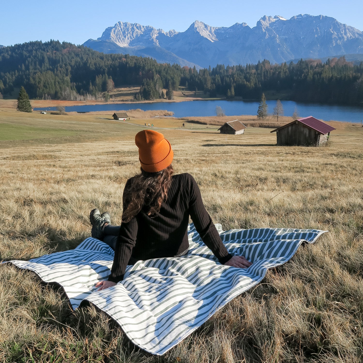 Person sitzt auf einer Decke auf einer Wiese und blickt auf einen See und Berge im Hintergrund.