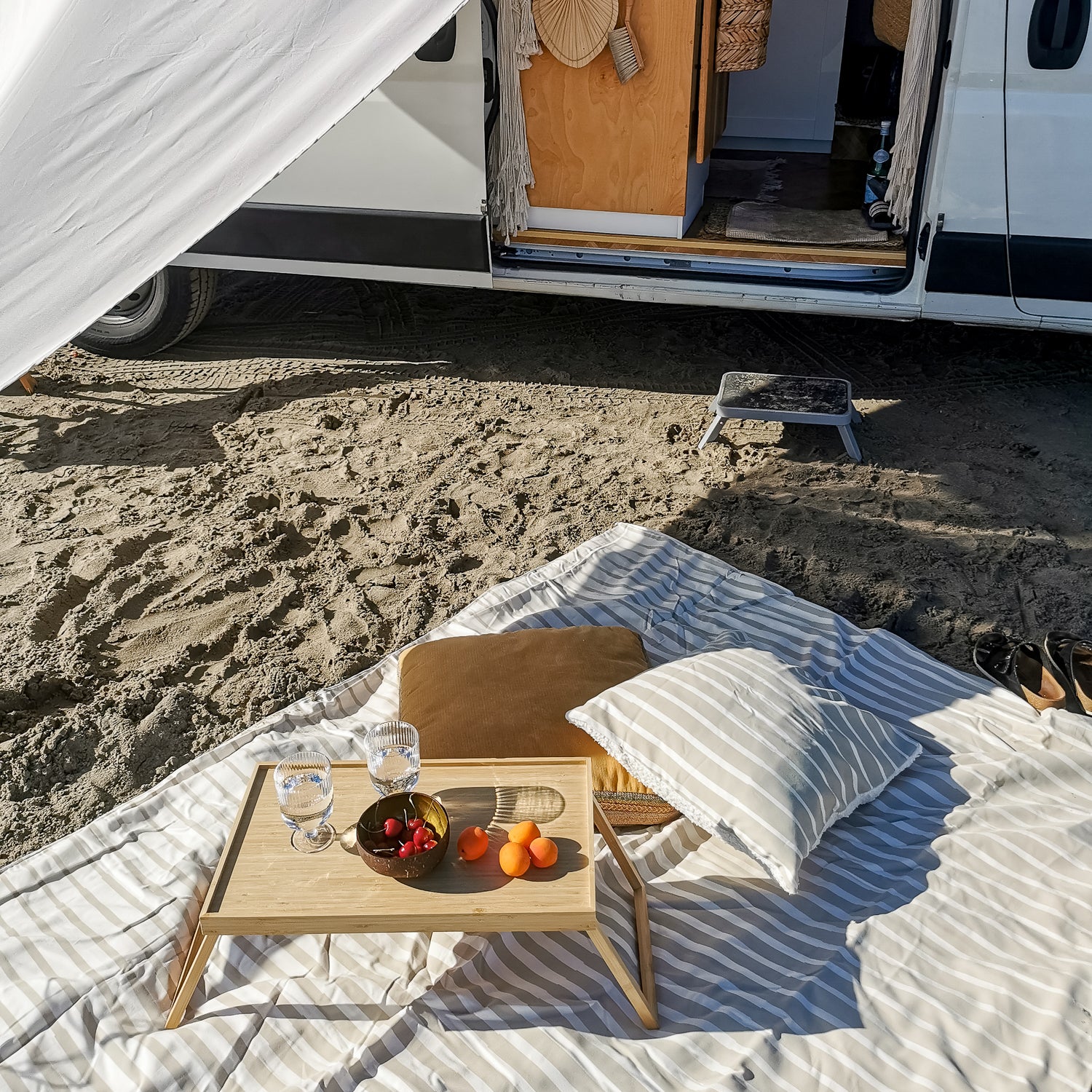 Picknick-Setup auf einem Streifen-Teppich vor einem offenen Van mit Getränken, Früchten, Kissen und Sandboden.
