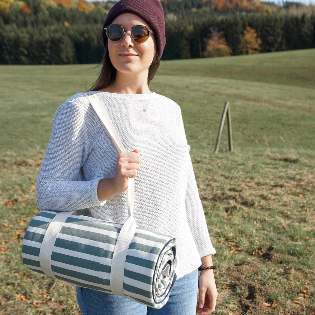 Frau mit Mütze und Sonnenbrille trägt eine gestreifte Decke über der Schulter auf einem Feld.