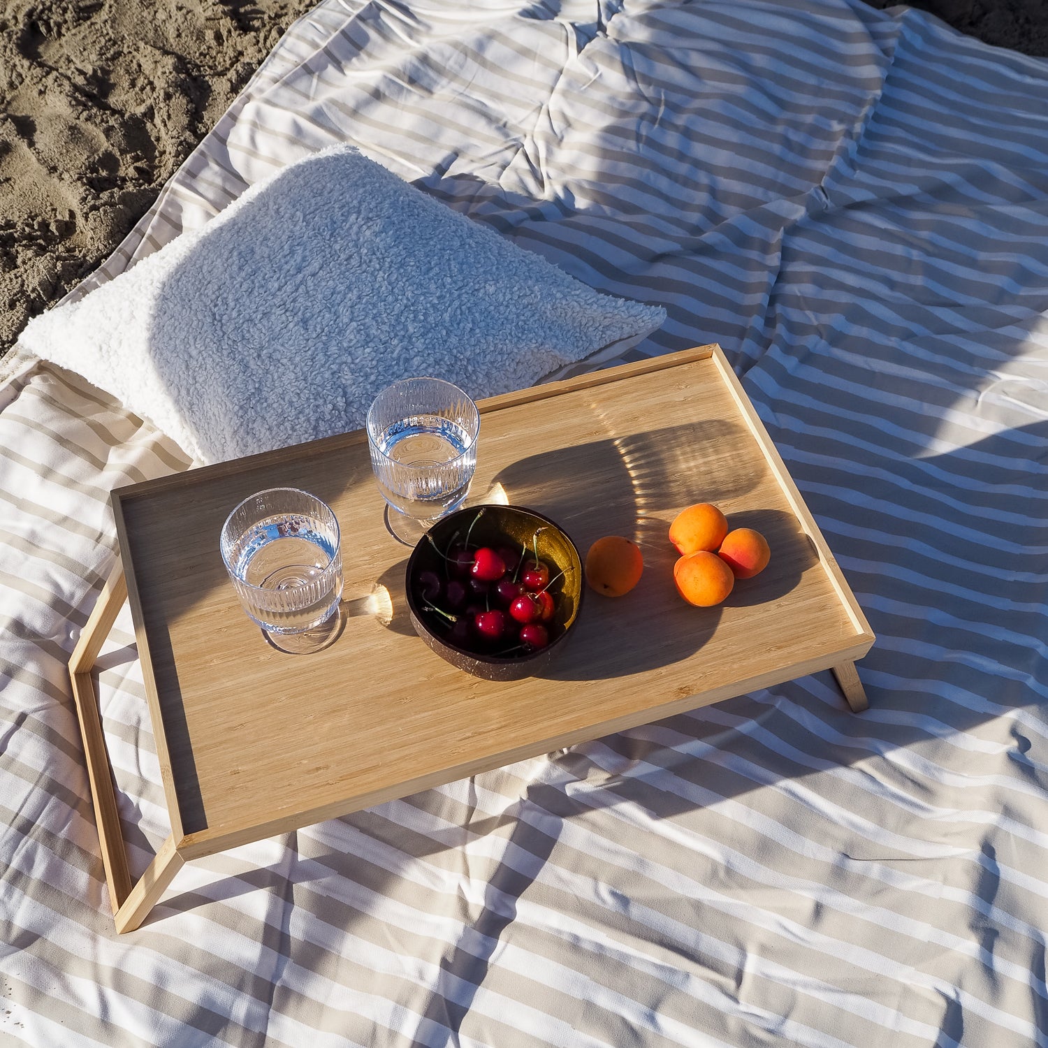 Holztisch auf gestreiftem Strandtuch mit zwei Wassergläsern, einer Schale Kirschen und Aprikosen, neben einem Kissen.