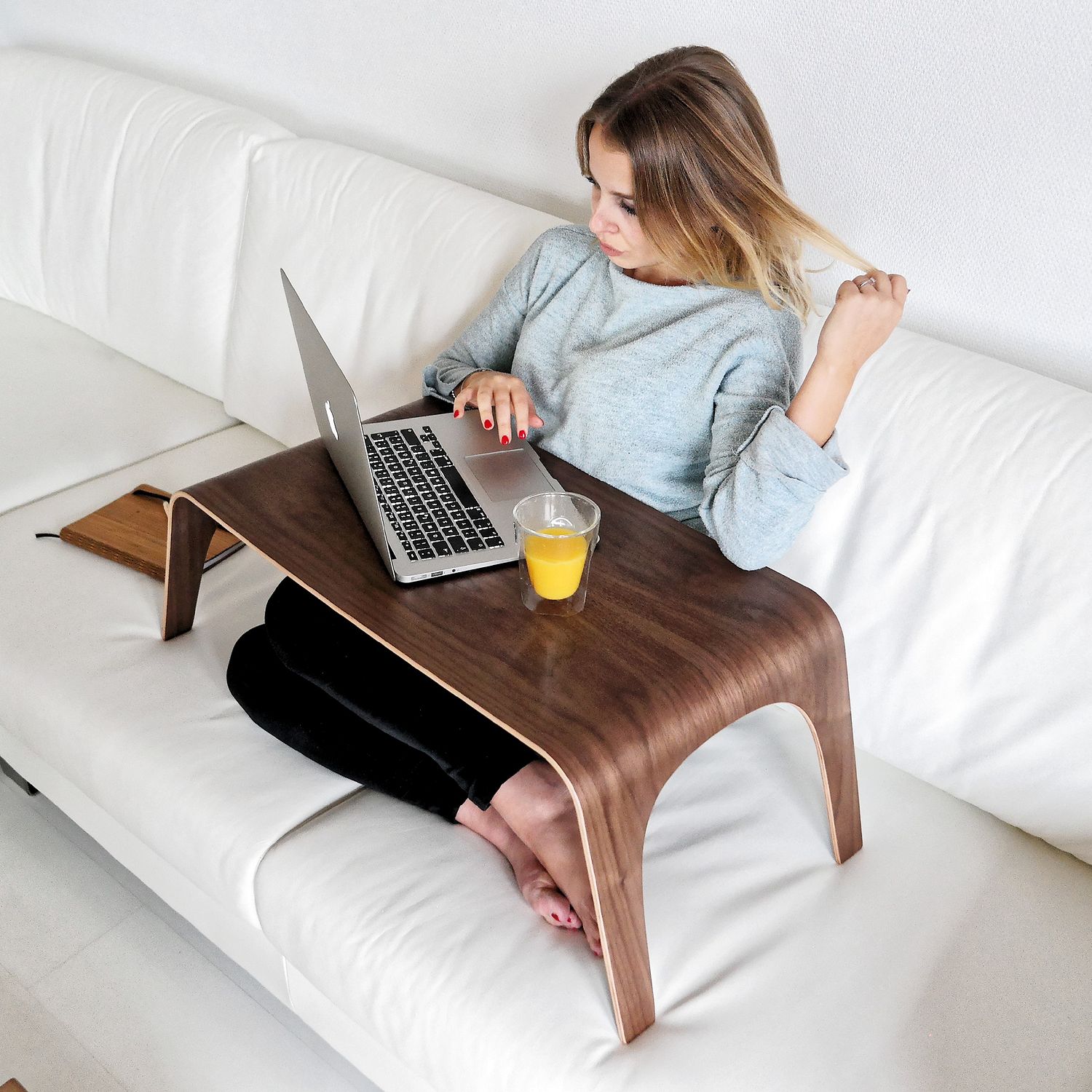 Frau sitzt auf einem Sofa mit Laptop auf einem Holztablett-Tisch und einem Glas Orangensaft.
