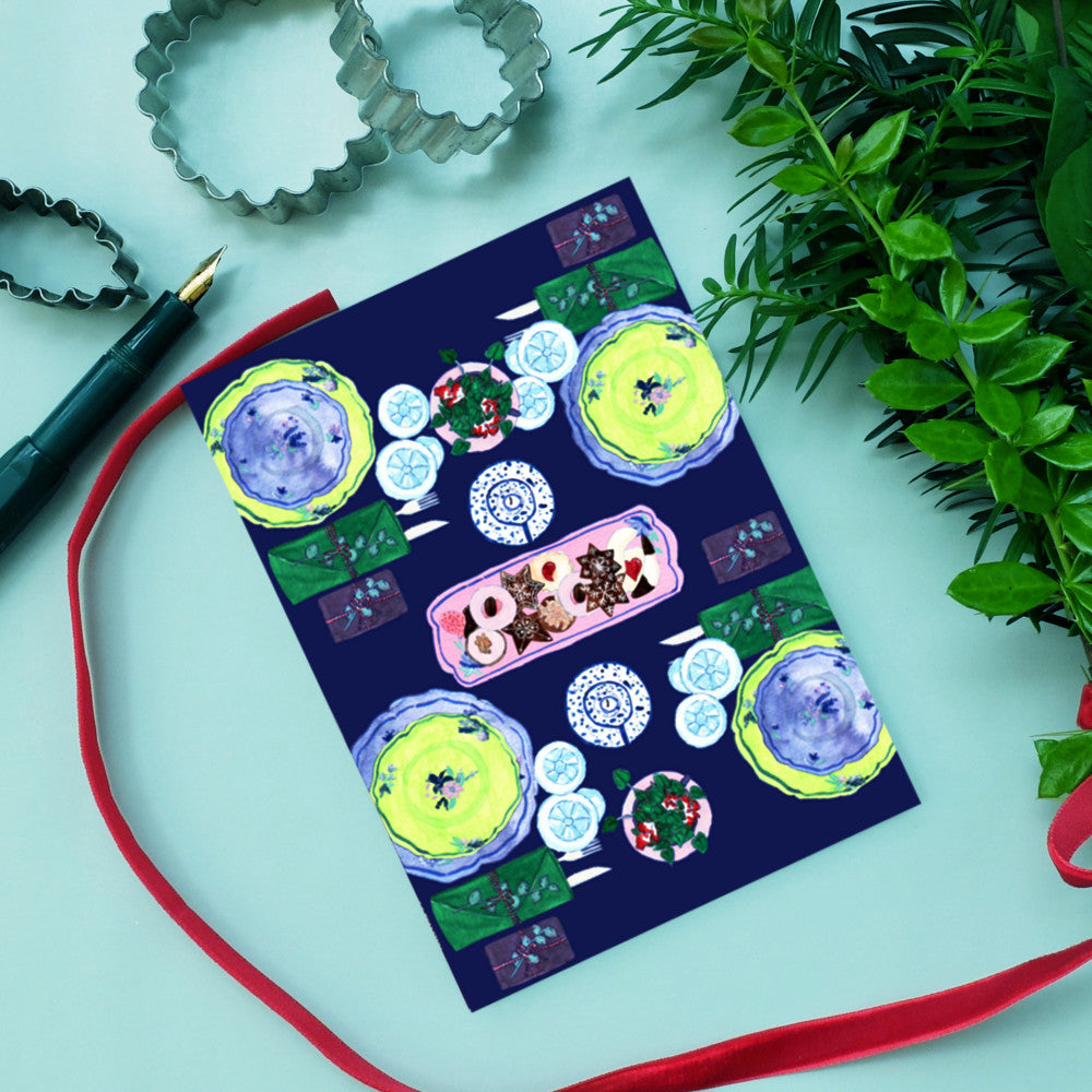 Eine Postkarte mit buntem, handgemaltem Tischgedeck, neben roten Bändern und grünen Blättern auf hellem Hintergrund.