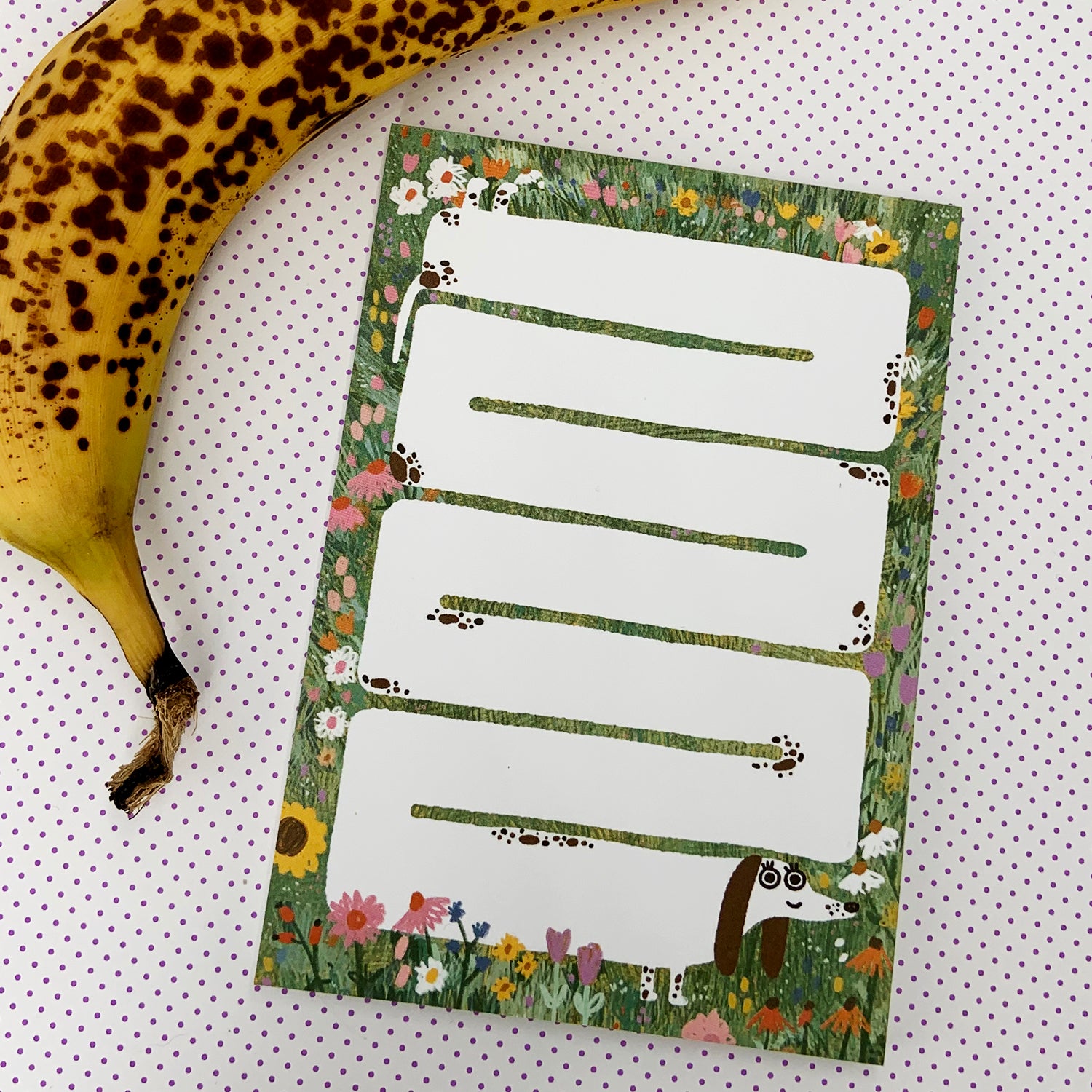 Eine braune reife Banane neben einer Karte mit weißen Etiketten und Blumenmuster auf gepunktetem Hintergrund.