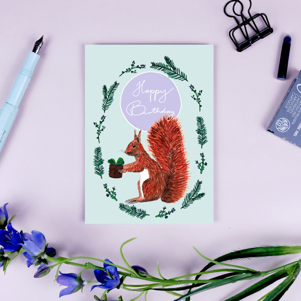Geburtstagskarte mit einem Eichhörnchen, das einen Blumentopf hält, umgeben von Tannenzweigen und dem Text "Happy Birthday".