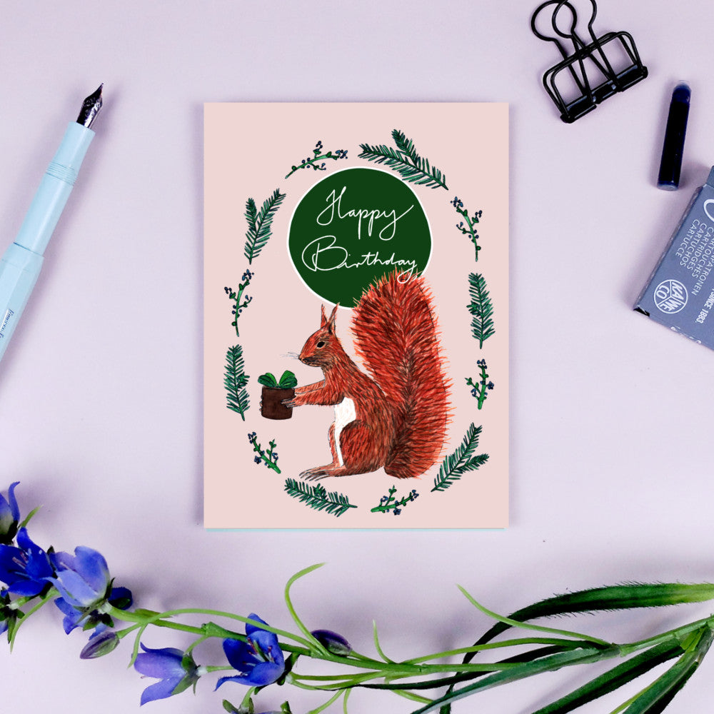 Geburtstagskarte mit einem Eichhörnchen, das einen Blumentopf hält, umgeben von Zweigen und dem Text "Happy Birthday".
