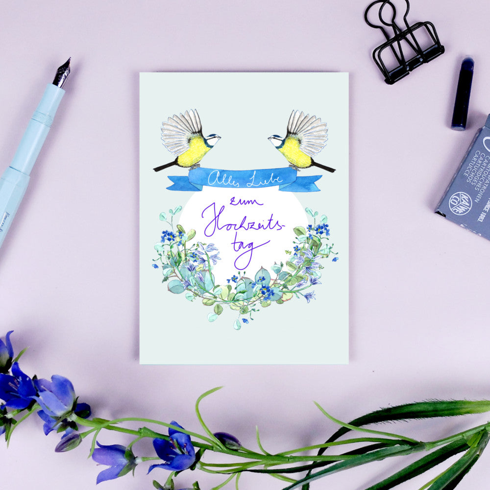 Hochzeitskarte mit zwei Vögeln, blauer Schleife und Blütenkranz, umgeben von Schreibutensilien und blauen Blumen.