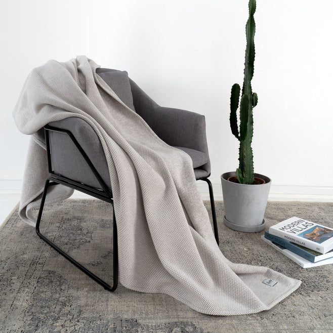 Grauer Sessel mit beiger Decke, daneben steht ein Kaktus in einem Topf und es liegen Bücher auf dem Boden.