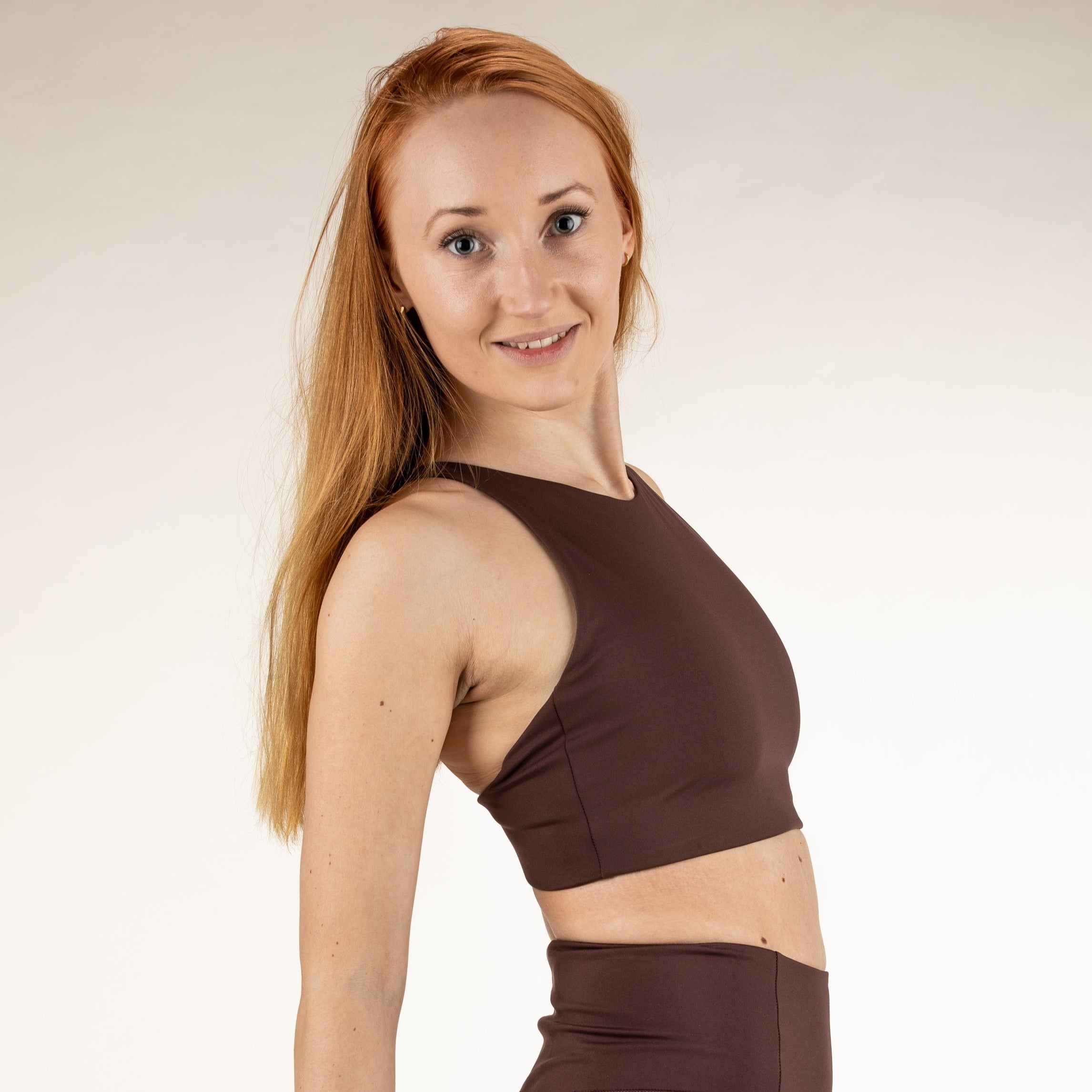 Frau mit langen roten Haaren lächelt in die Kamera und trägt ein braunes Sportoberteil und Leggings.