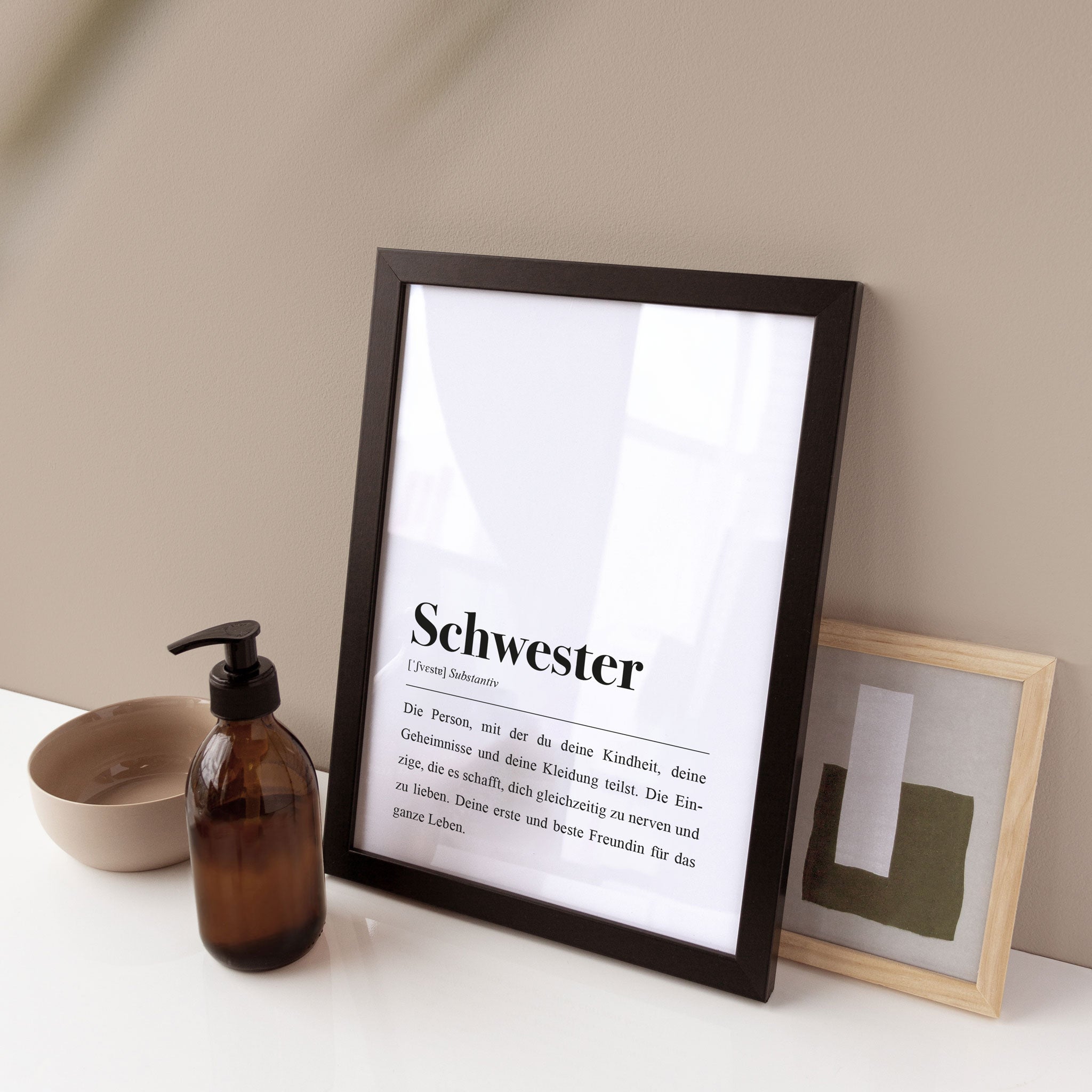 Gerahmtes Bild mit Definition von "Schwester" neben einer braunen Glasspenderflasche und einem kleinen Kunstwerk.