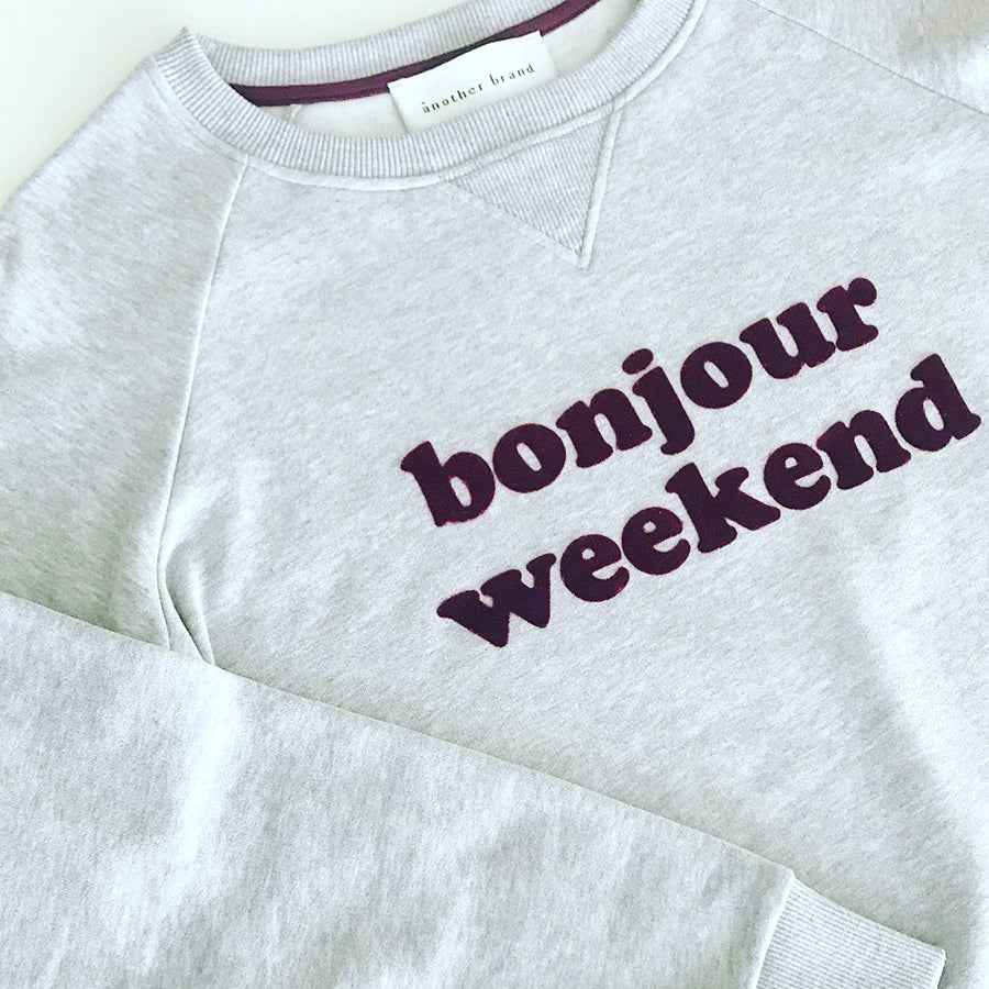 Grauer Pullover mit lila Schriftzug "bonjour weekend" auf der Vorderseite, Label "another brand" im Nackenbereich.