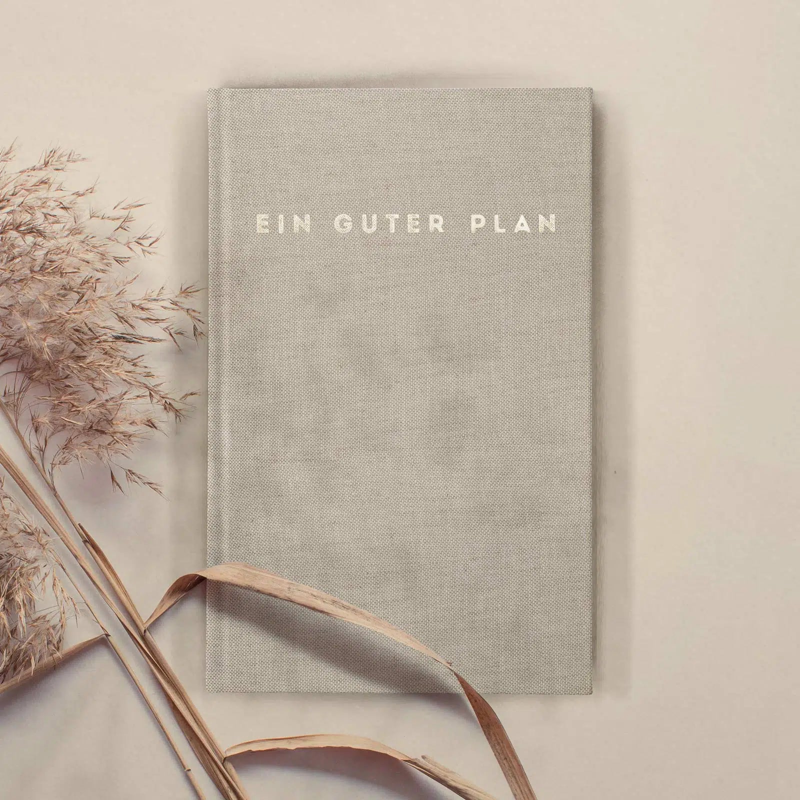Graues Notizbuch mit der Aufschrift "Ein Guter Plan", daneben getrocknete Gräser auf beigem Hintergrund.