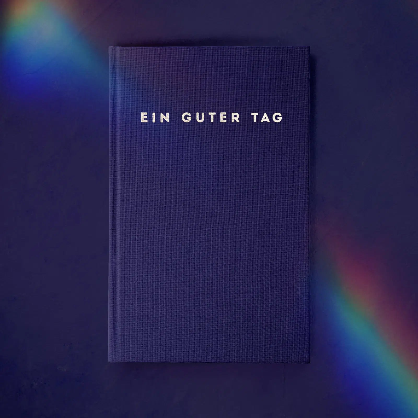 Ein blaues Buch mit der Aufschrift "Ein Guter Tag", umgeben von einem schwachen Regenbogen auf dunklem Hintergrund.