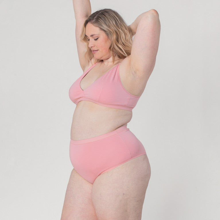 Eine Person in rosa Unterwäsche hebt beide Arme vor einem neutralen Hintergrund.