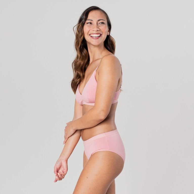 Eine lächelnde Frau trägt ein rosa Unterwäsche-Set vor einem neutralen Hintergrund.