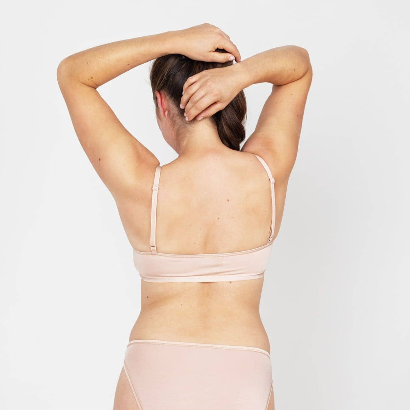 Frau in beigefarbenem Unterwäsche-Set, von hinten fotografiert, hebt die Arme über den Kopf vor weißem Hintergrund.