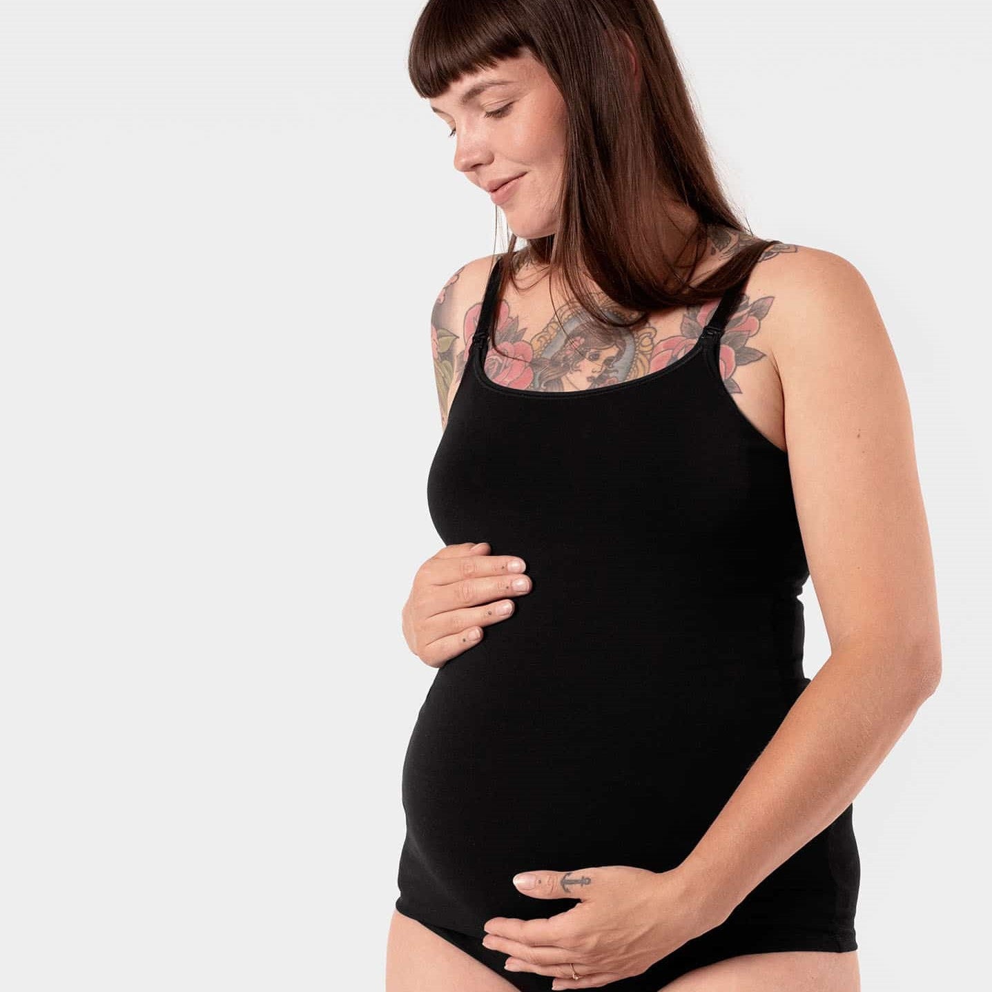 Schwangere Frau in schwarzem Tanktop mit Tattoos, die sanft ihren Bauch hält, auf weißem Hintergrund.