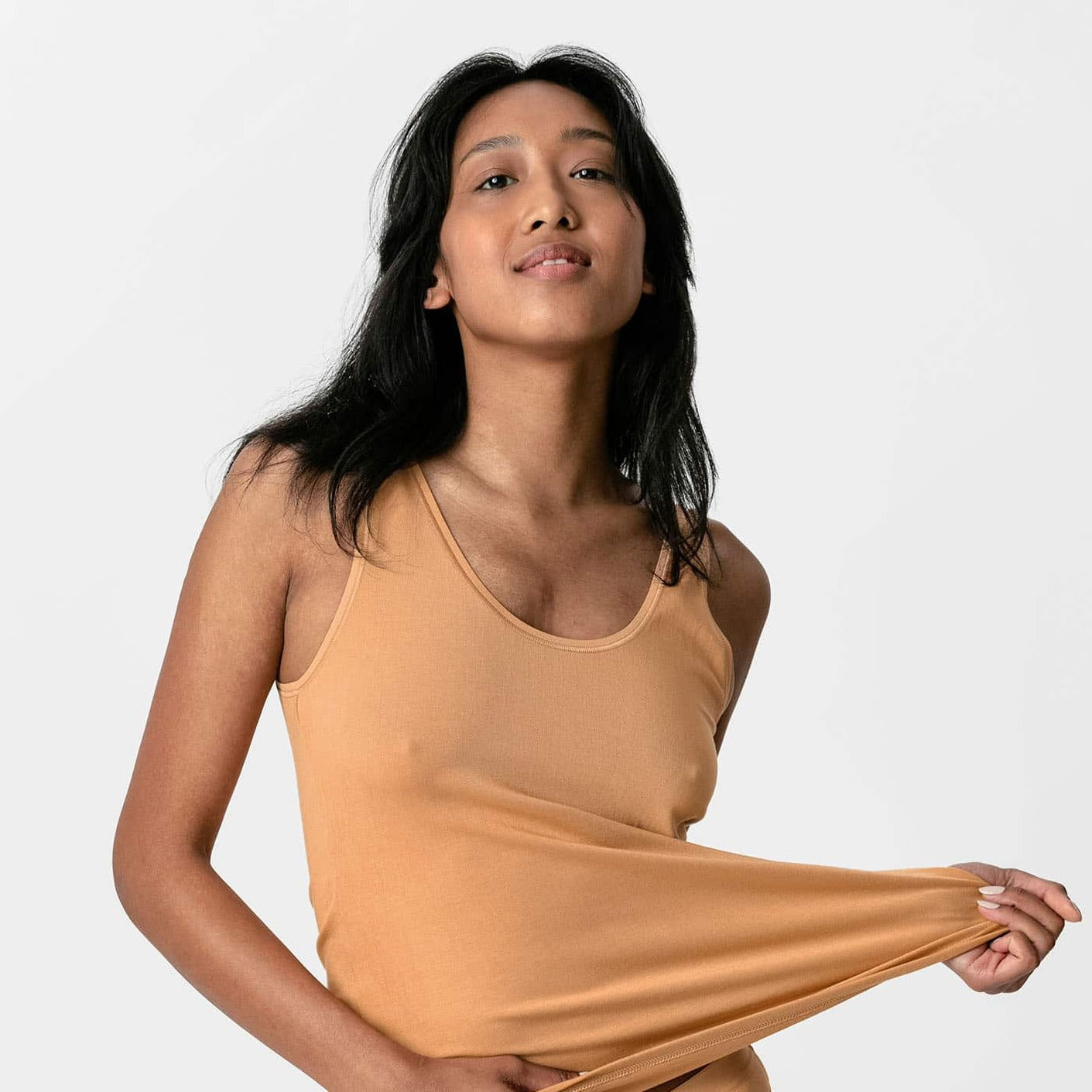 Person in beige tank top holds its hem, posing confidently against a plain background.